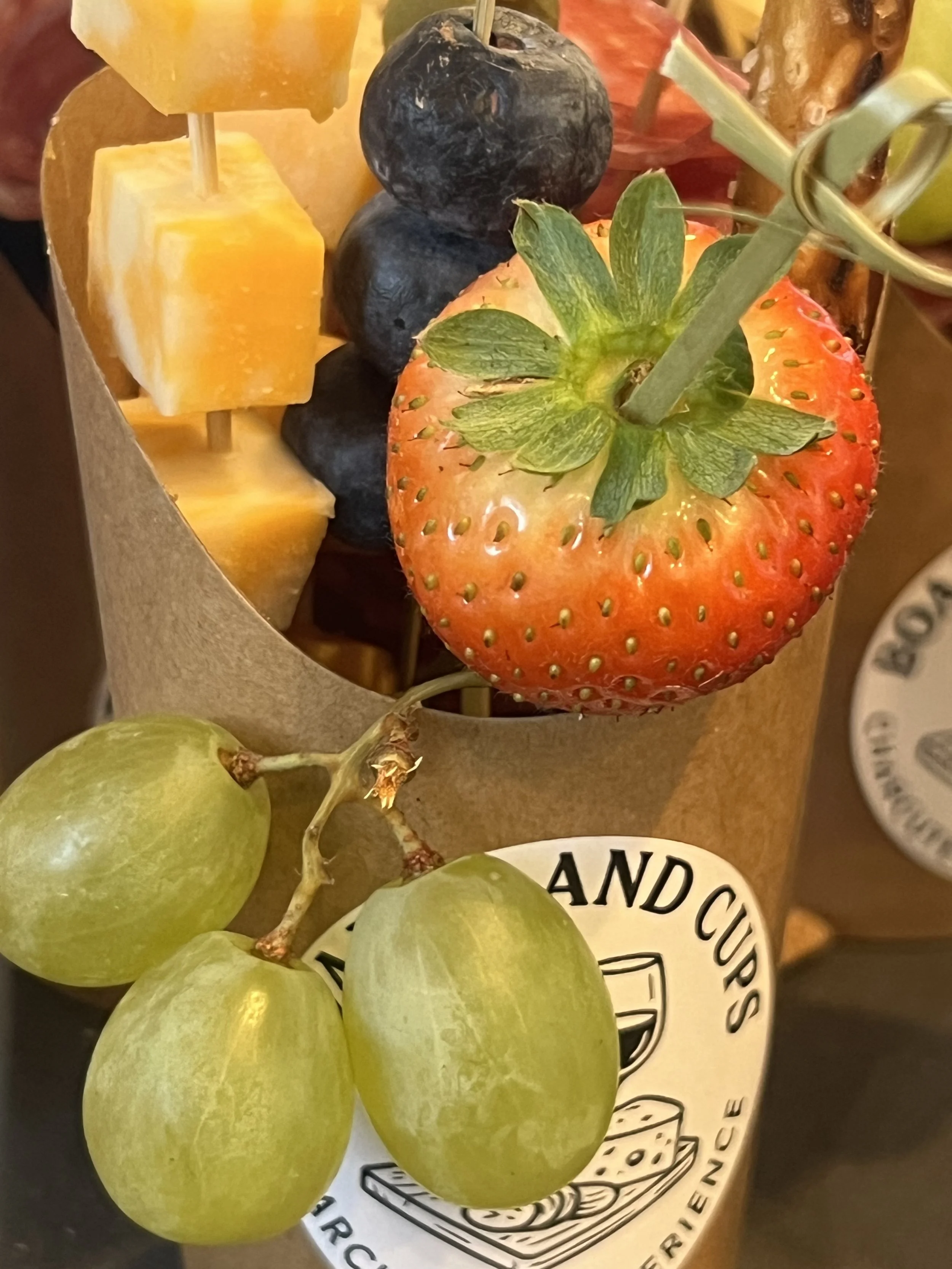 A bouquet with skewered cheese, black blueberries, a large strawberry, and green grapes, wrapped in brown paper with a label.