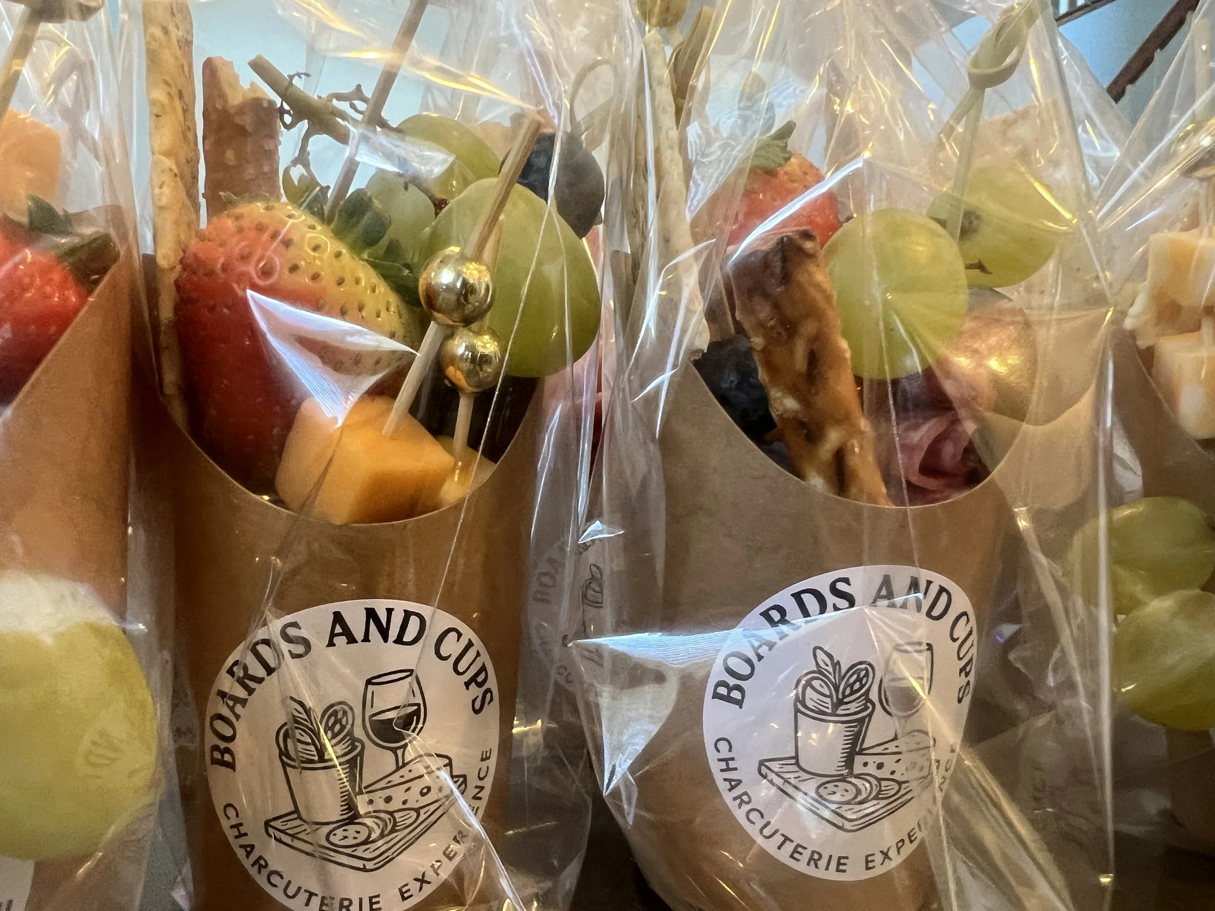 Clear plastic bags containing assorted cut fruits such as strawberries, green grapes, honeydew, cantaloupe, and pineapple, with some fruit skewered on sticks and decorated with small metallic silver balls. The bags are labeled with a logo reading 'Boards and Cups Charcuterie Experience'.