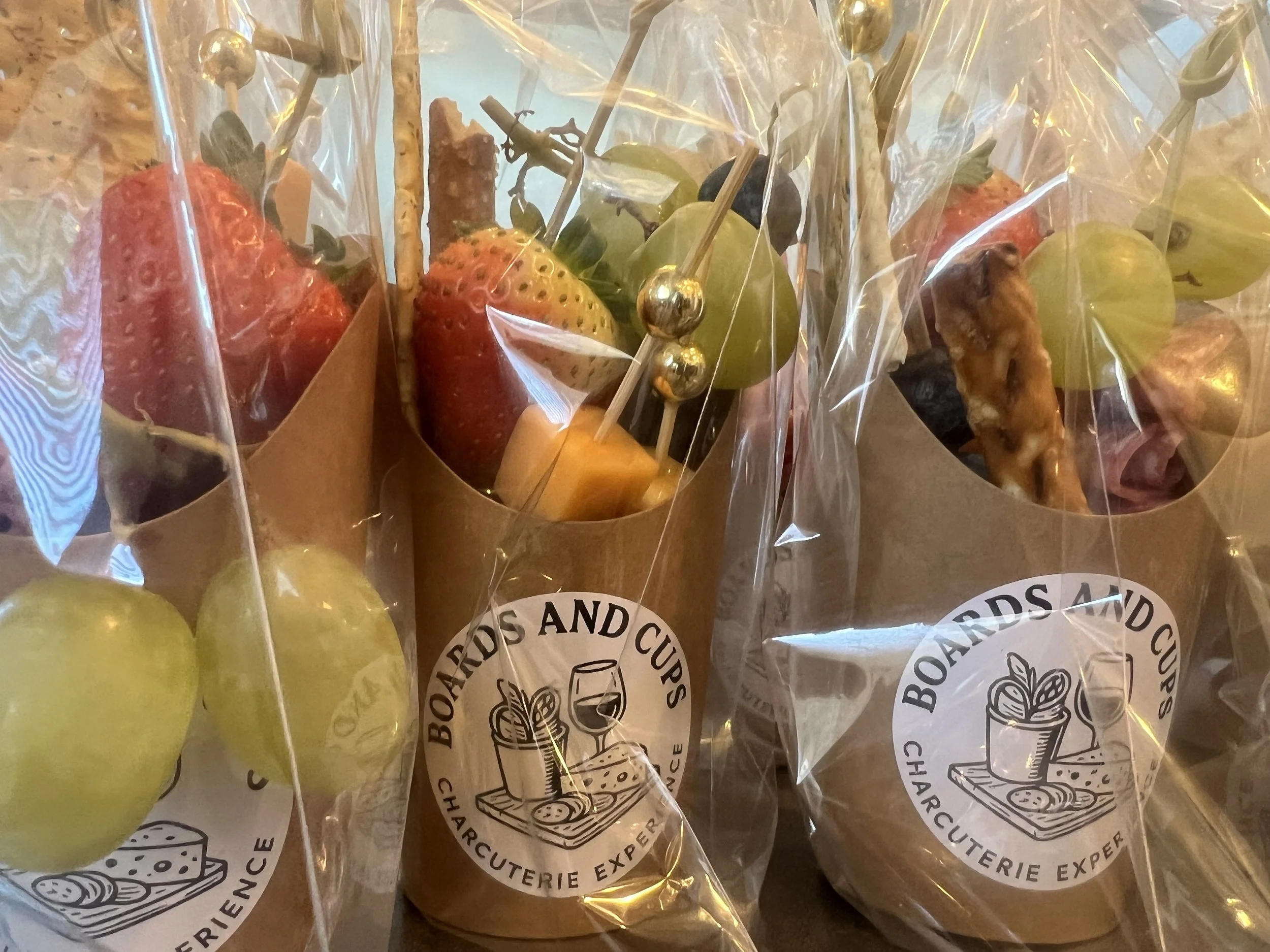 Small fruit bouquets wrapped in clear plastic with a label reading 'Boards and Cups Charcuterie Expert.' The bouquets contain various fruits, including strawberries, green grapes, and cheese cubes, with some skewers also holding nuts or crackers.