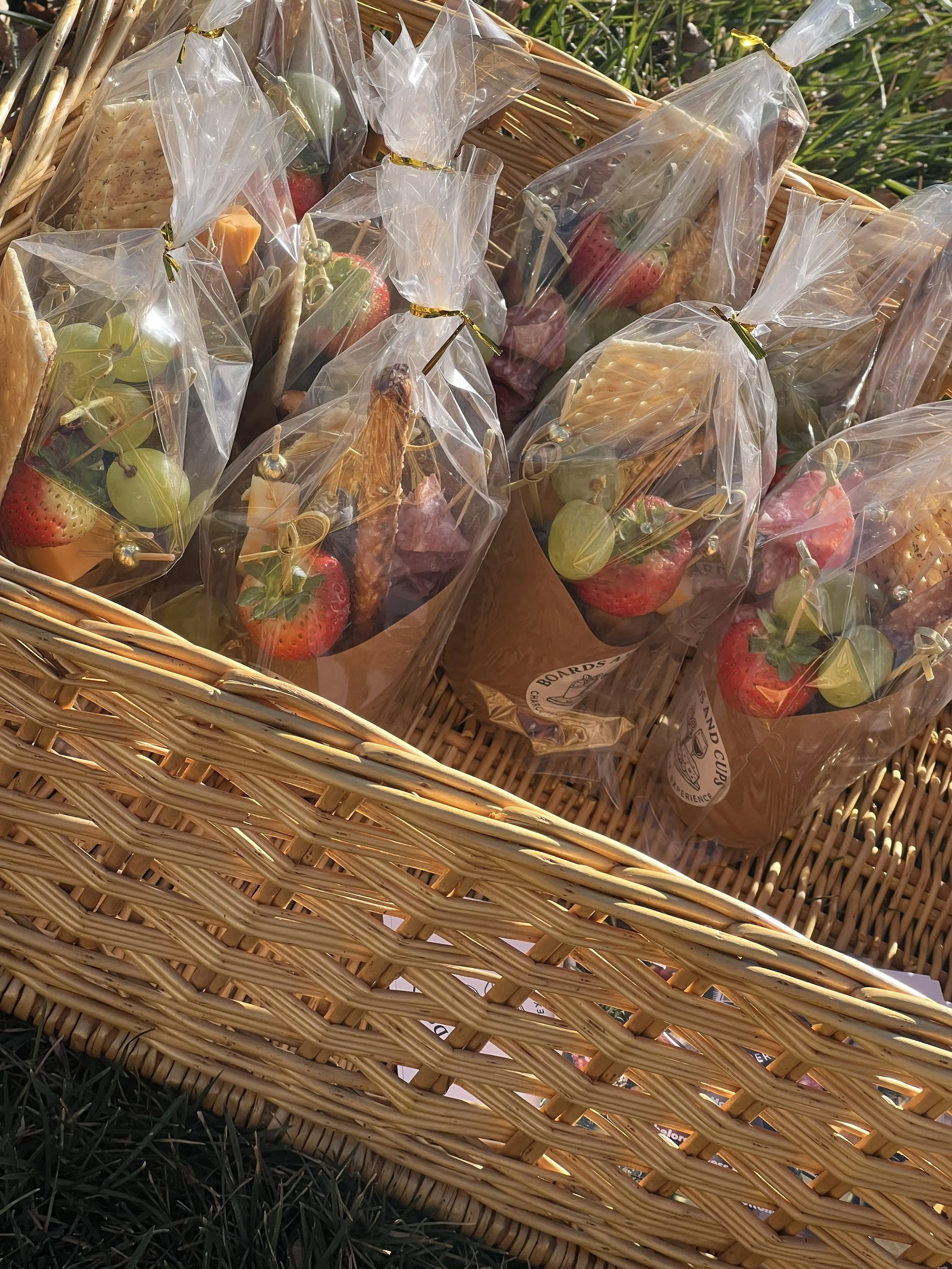 Small plant pots with strawberries and green grapes, wrapped in clear plastic and tied with gold twist ties, inside a wicker basket.