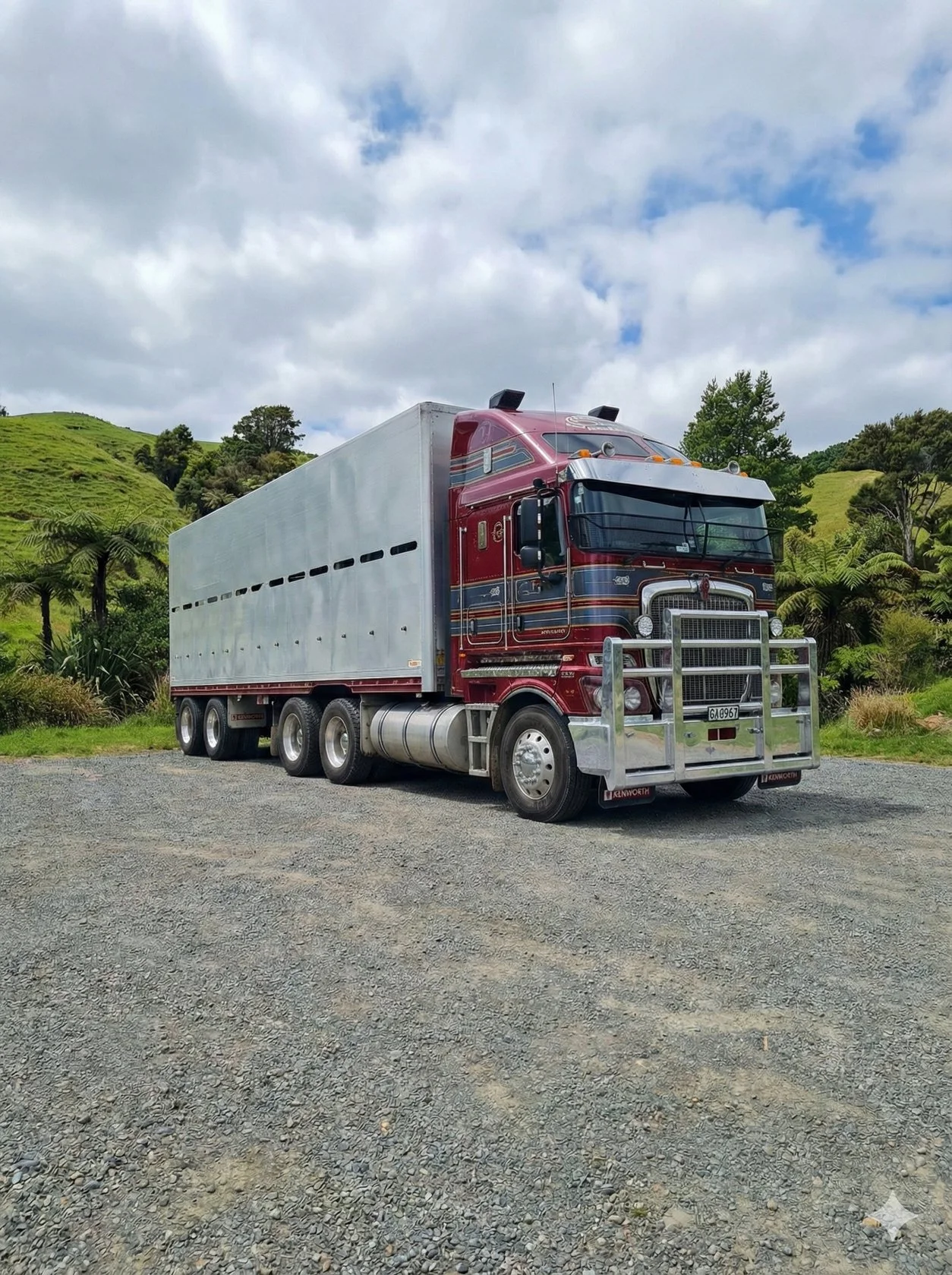 A large semi-truck with a shiny red cab and a gray trailer parked on a gravel lot with green hills and trees in the background, under a partly cloudy sky.