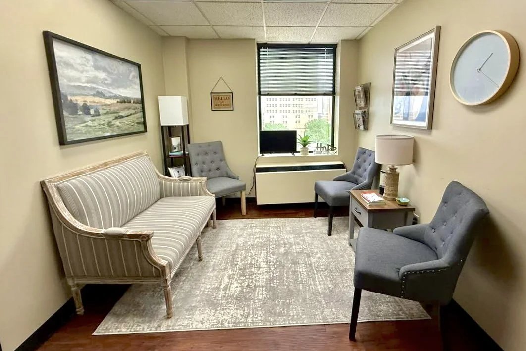 A small waiting room with four chairs, a side table, a lamp, a wall clock, a record of framed pictures, a window, and a painting of a landscape.