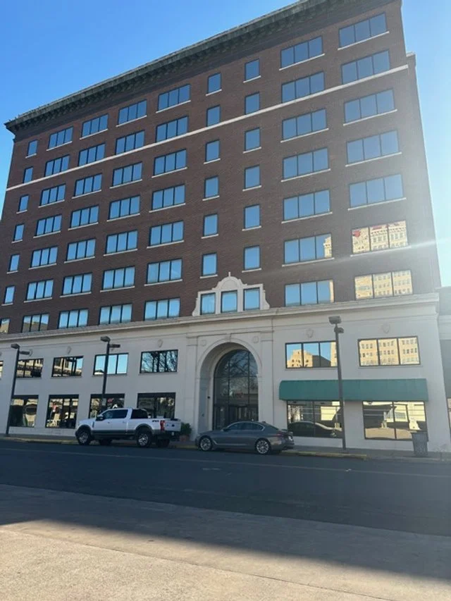 A tall, multi-story brick building with large windows on each floor. The ground floor has a white ornate entrance and a storefront with green awnings. Several cars are parked in front, and the sky is clear and blue.