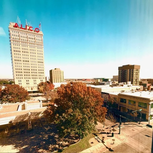 Cityscape with a tall building bearing a red ALCO sign, trees with fall foliage, and lower buildings under a clear blue sky.