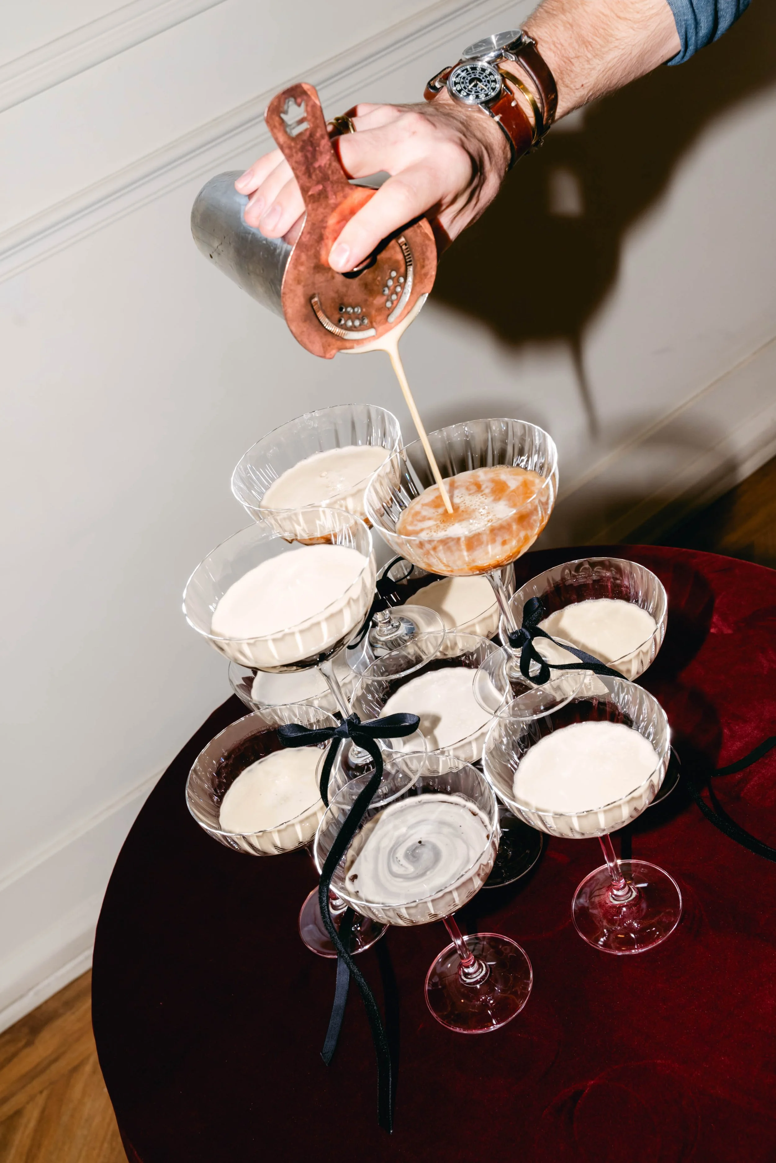 Person pouring a layered cocktail from a copper shaker into a coupe glass, surrounded by other filled coupe glasses on a table.