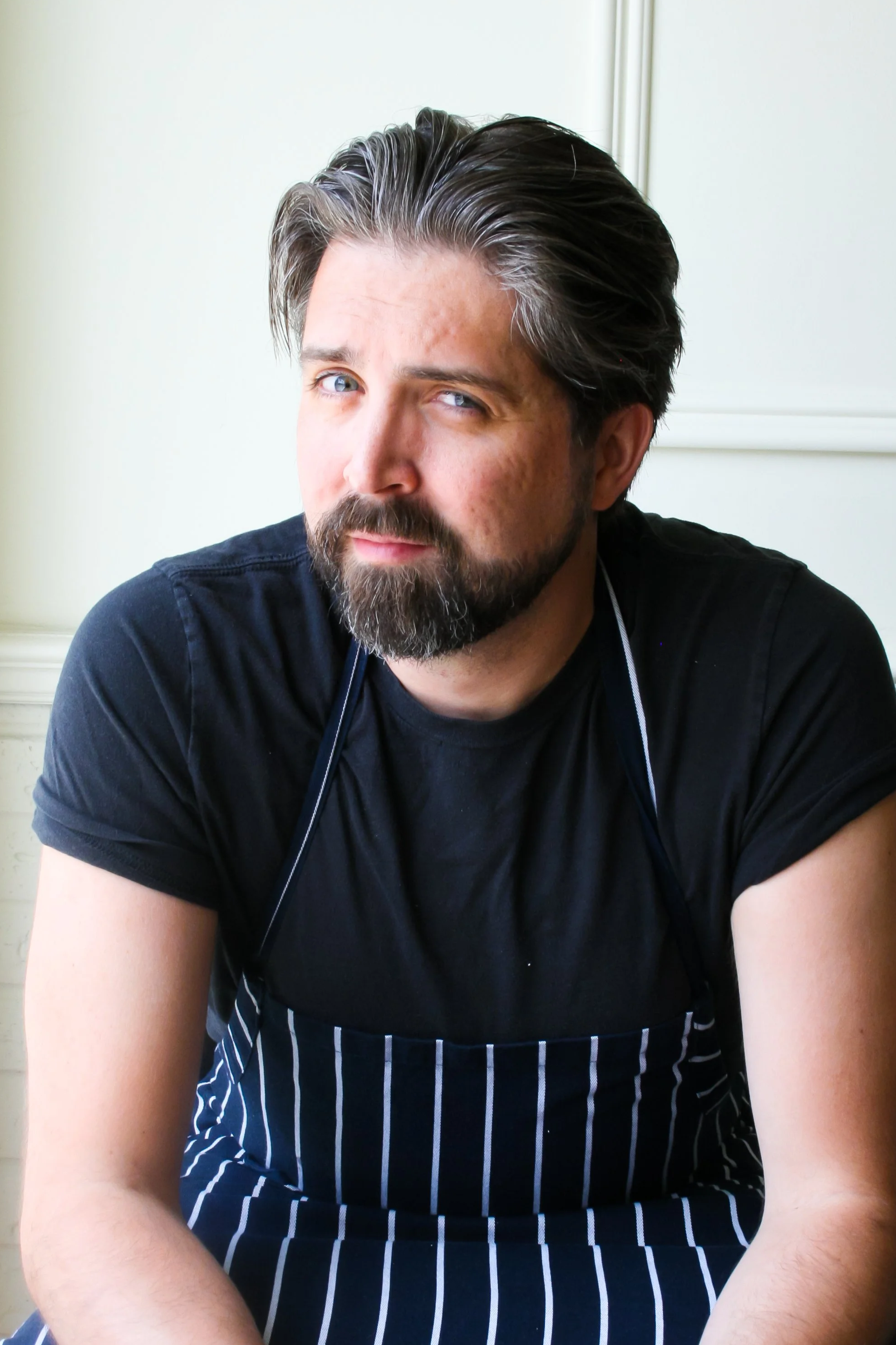 Andrew Ashpole, executive Chef and co-owner of Victoria's, with a beard and dark hair, wearing a black shirt and striped apron.