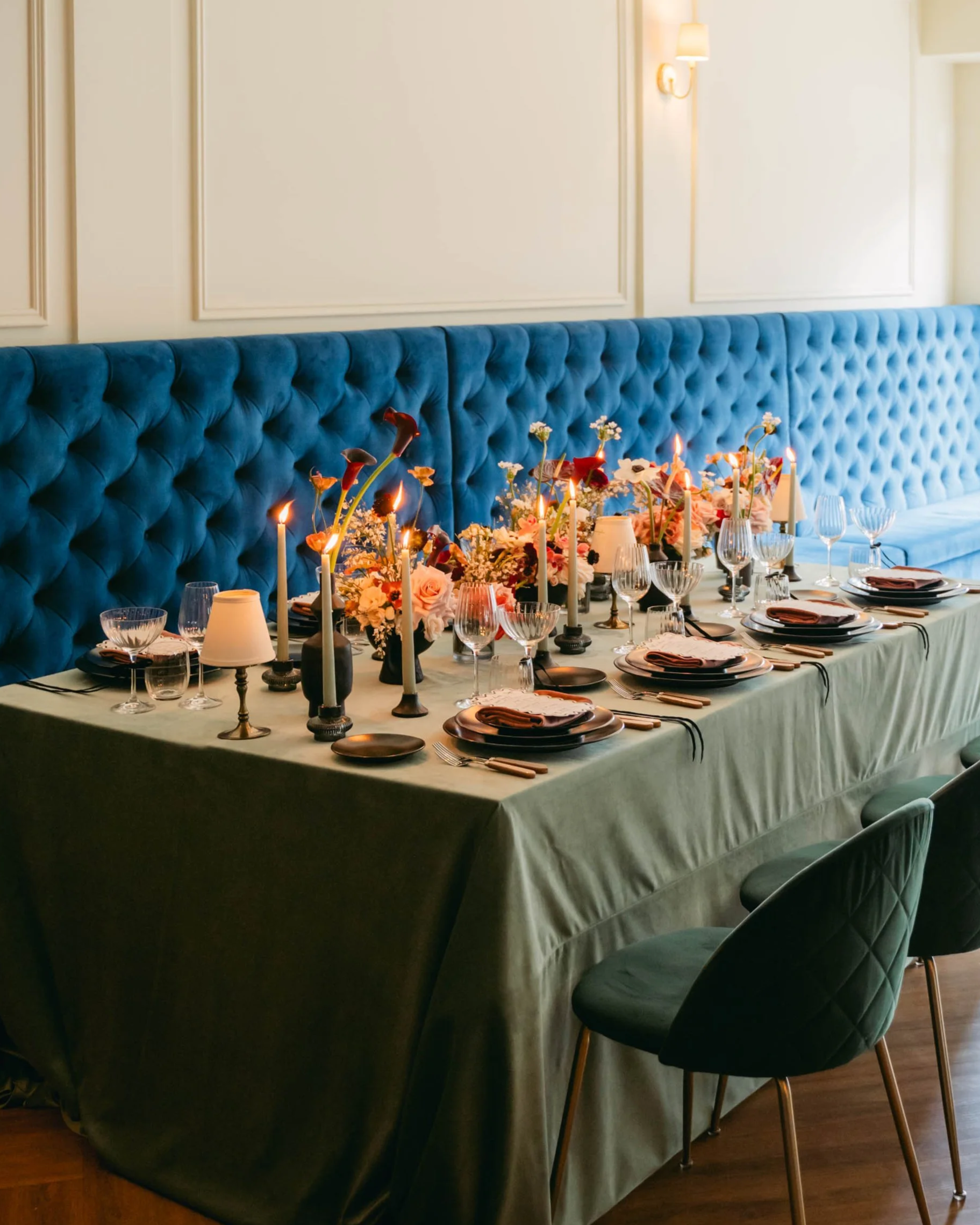 Elegant and modern dining table set for a wedding or celebration with floral centrepieces and dark tapered candles, against a blue tufted banquette.