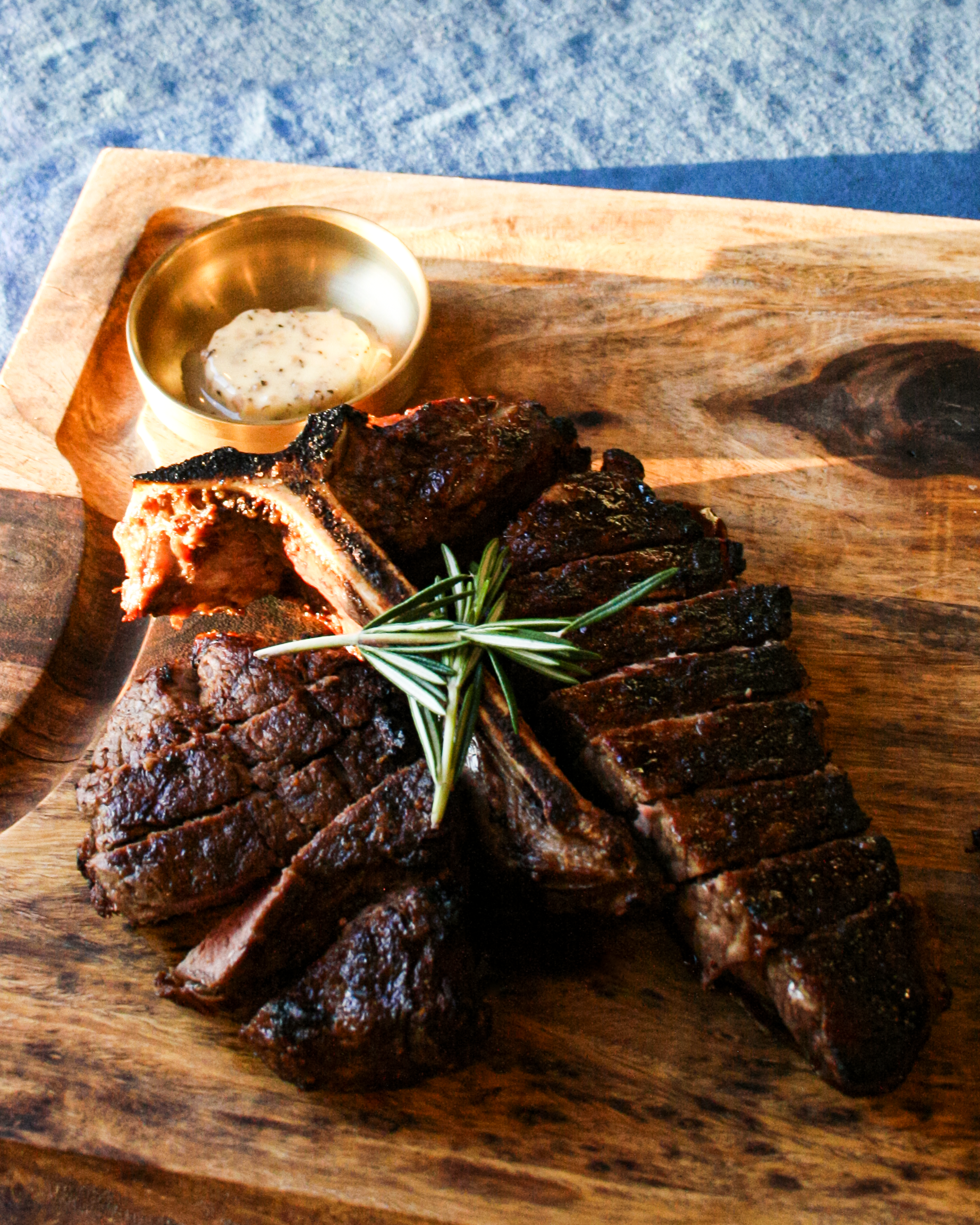 Bone-in Ribeye with a sprig of rosemary, served with a side of sauce on a wooden cutting board.