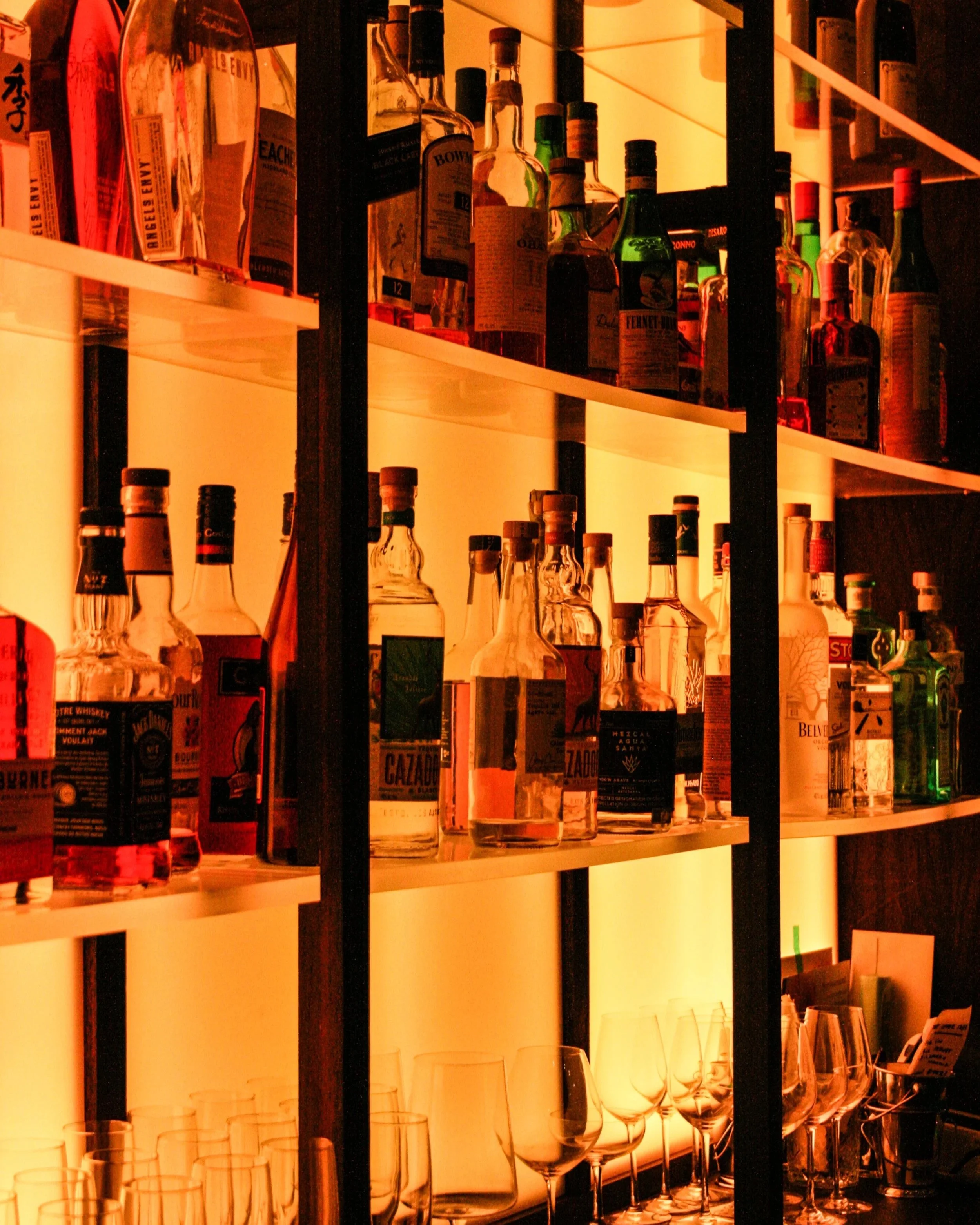 Shelves with various liquor bottles and glasses in a bar setting, illuminated with warm backlight.