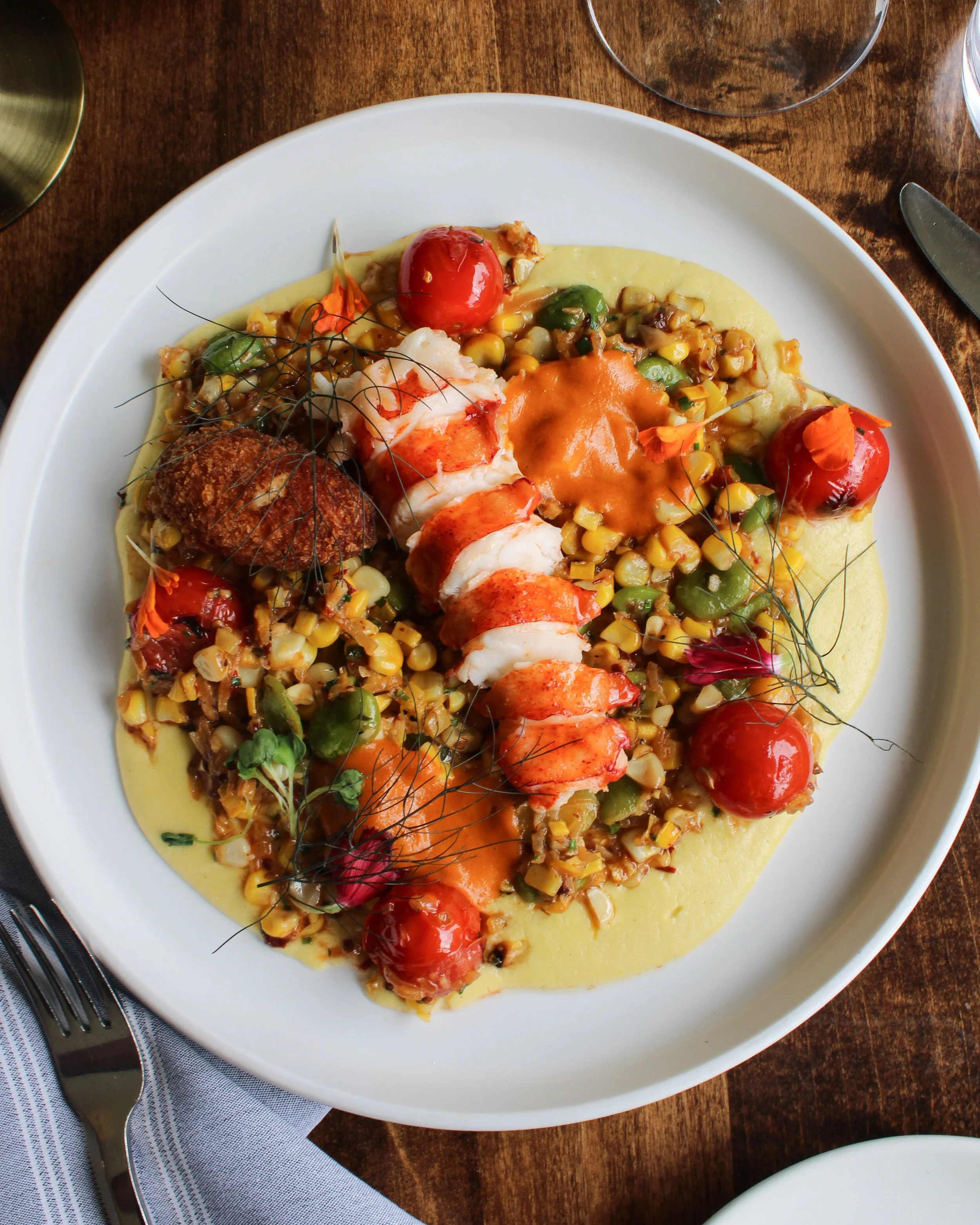 A colourful dish with succulent lobster tail, succotash, cherry tomatoes, microgreens, on a white plate.