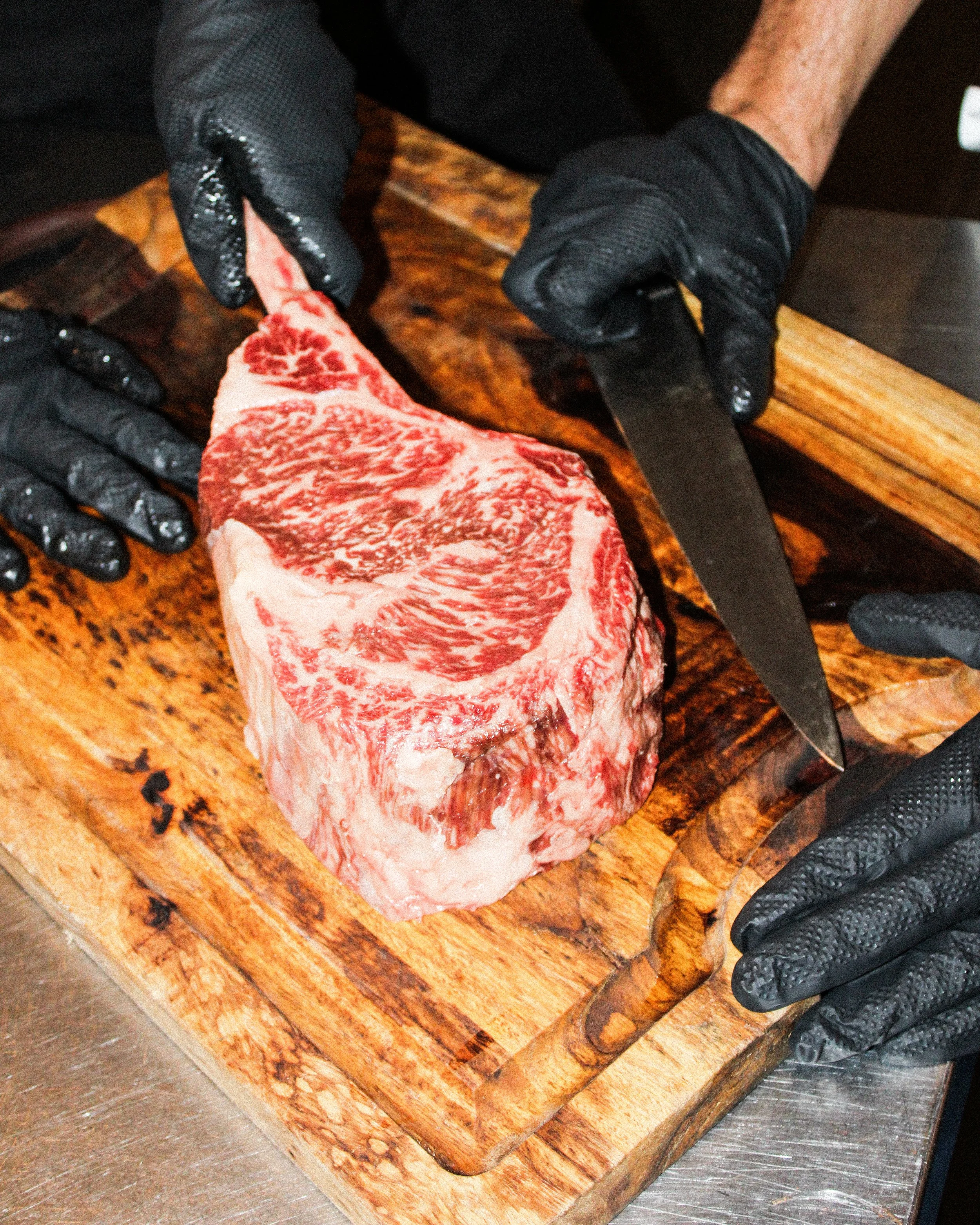 A person wearing black gloves is slicing a large, marbled cut of raw beef on a wooden cutting board with a large knife.