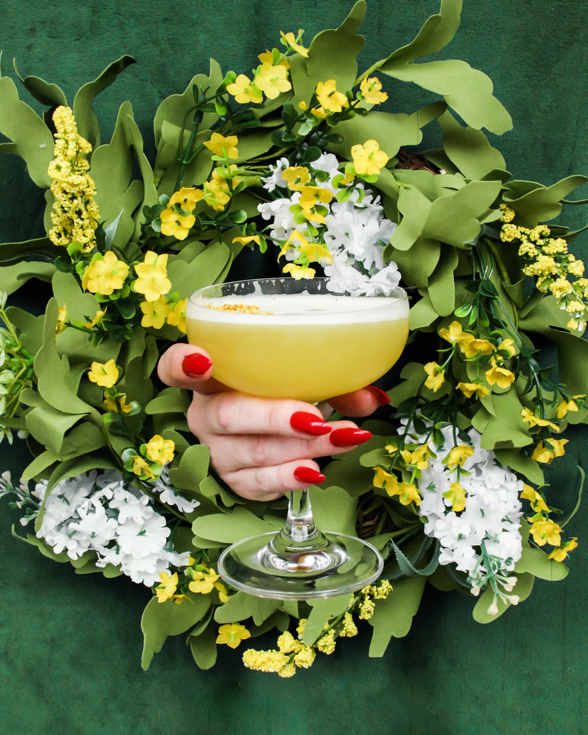 A very editorial, styled image featuring a hand with red painted nails holding a coupe with a yellow cocktail inside, surrounded by a green and yellow floral wreath and a green background.