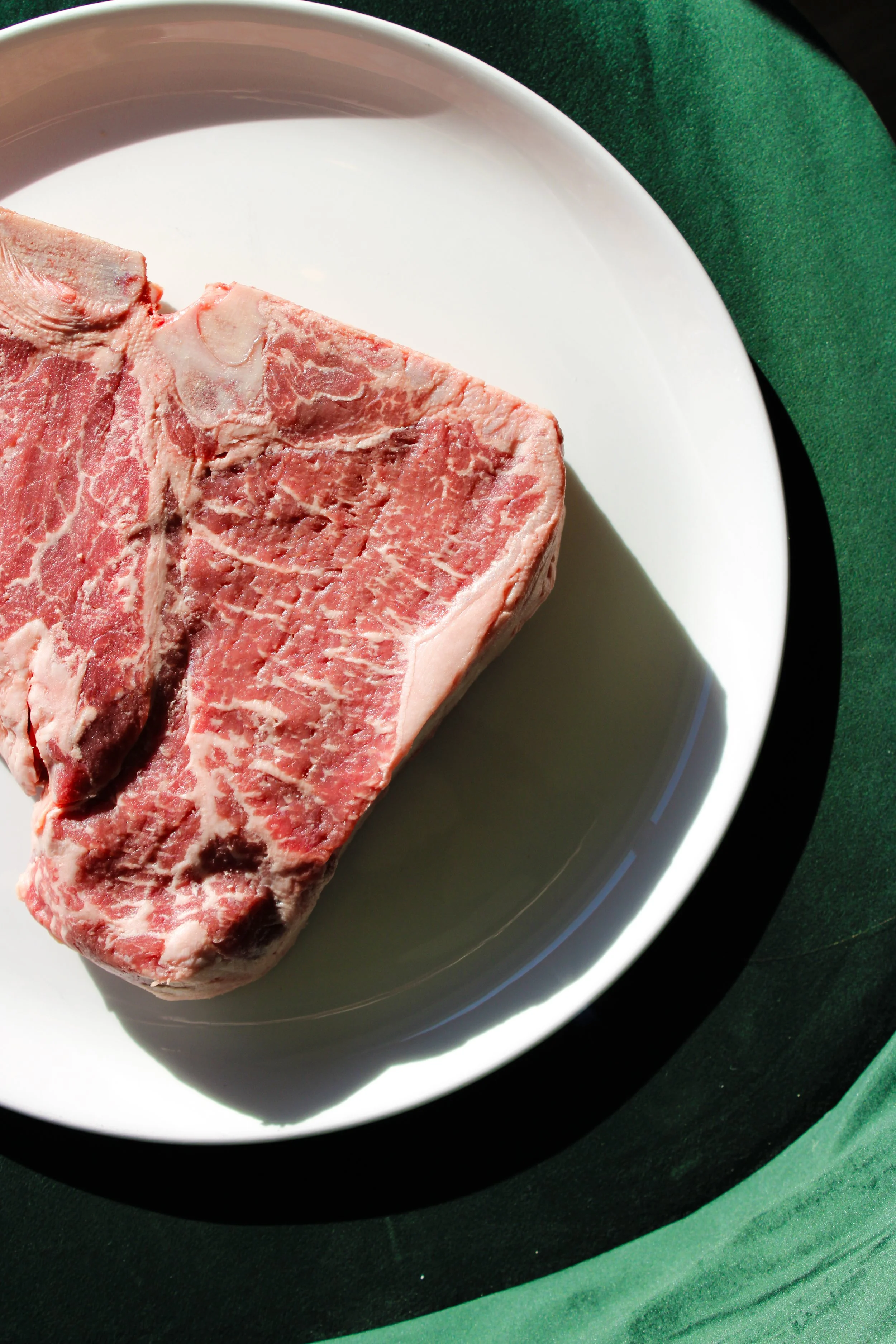 Raw T-bone steak on a white plate with green fabric underneath.