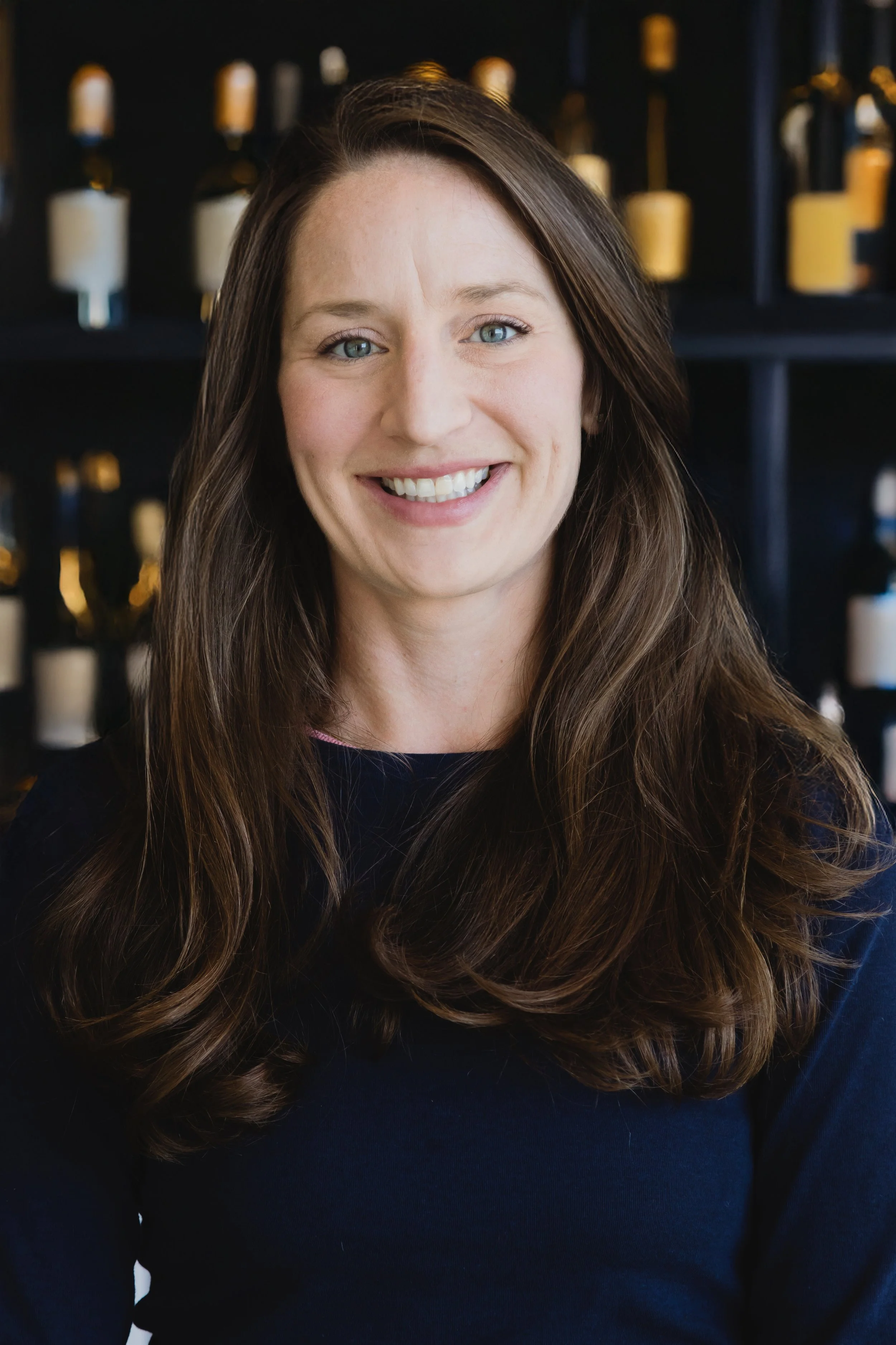Lois Connor, General Manager & co-owner of Victoria's, with long brown hair wearing a dark top, smiling, with a wine rack in the background.