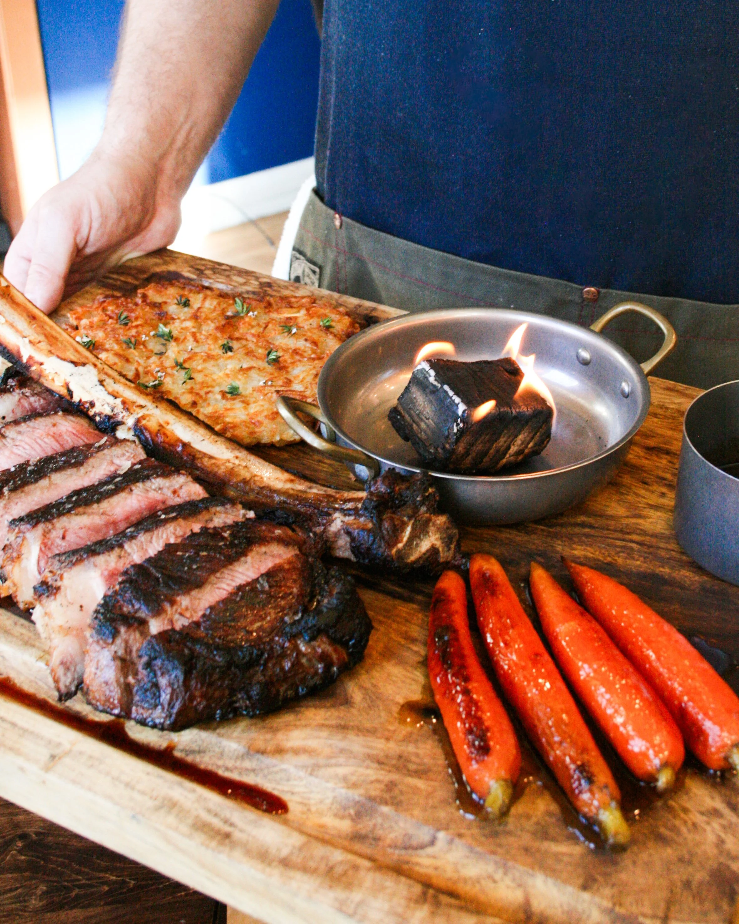 A person holding a large wooden cutting board with sliced medium-rare steak, roasted carrots, and a small skillet with a piece of burning wood, on a wooden table.