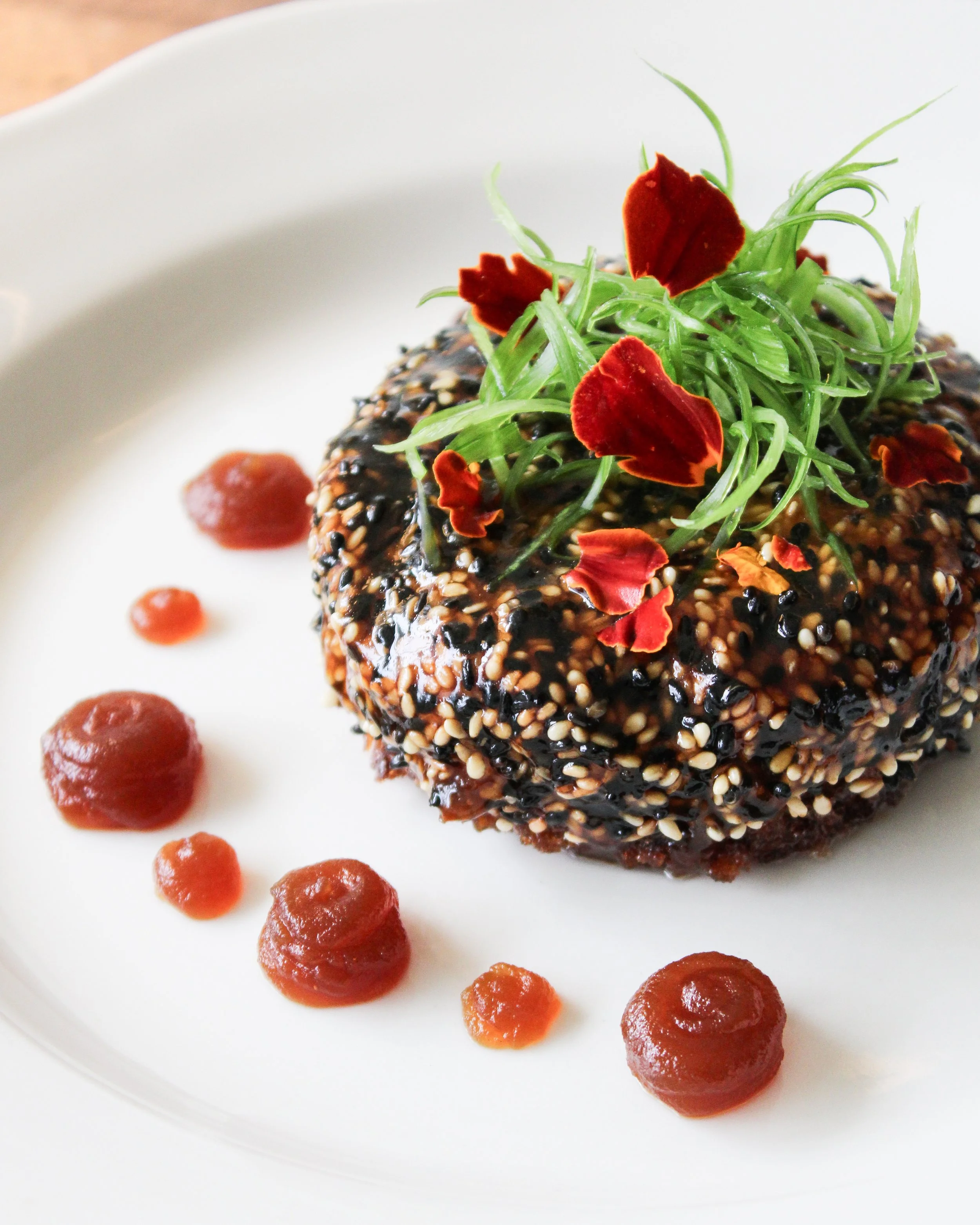 A round Scallop & Foie Toast featuring Japanese scallops, foie gras torchon, milk bread, sesame topped with microgreens and red flower petals, served with dots of red sauce on a white plate.
