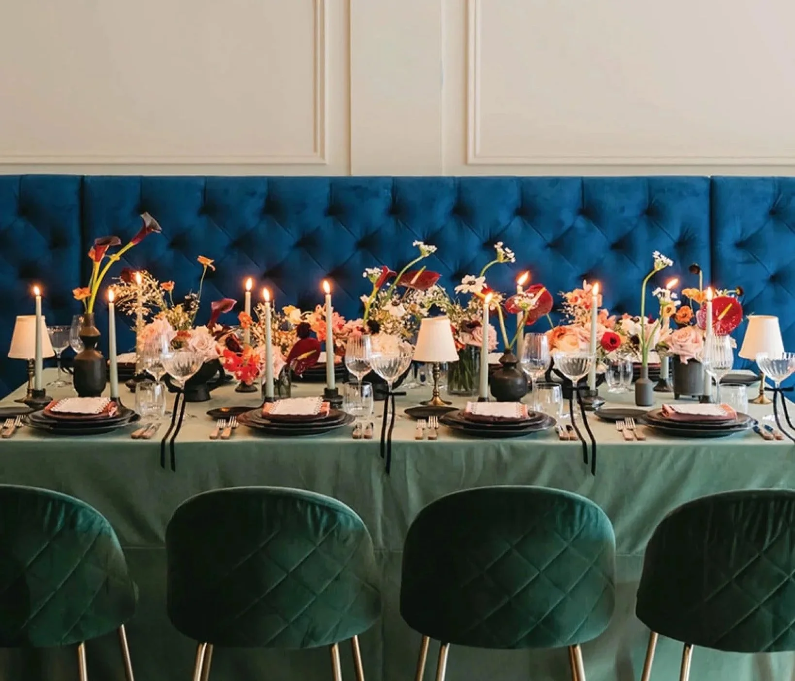 A formal dining table set for a modern restaurant Wedding in Hamilton, Ontario, decorated with pink and red flowers,, candles, black and white tableware, and green velvet chairs, with a blue tufted banquette in the background.