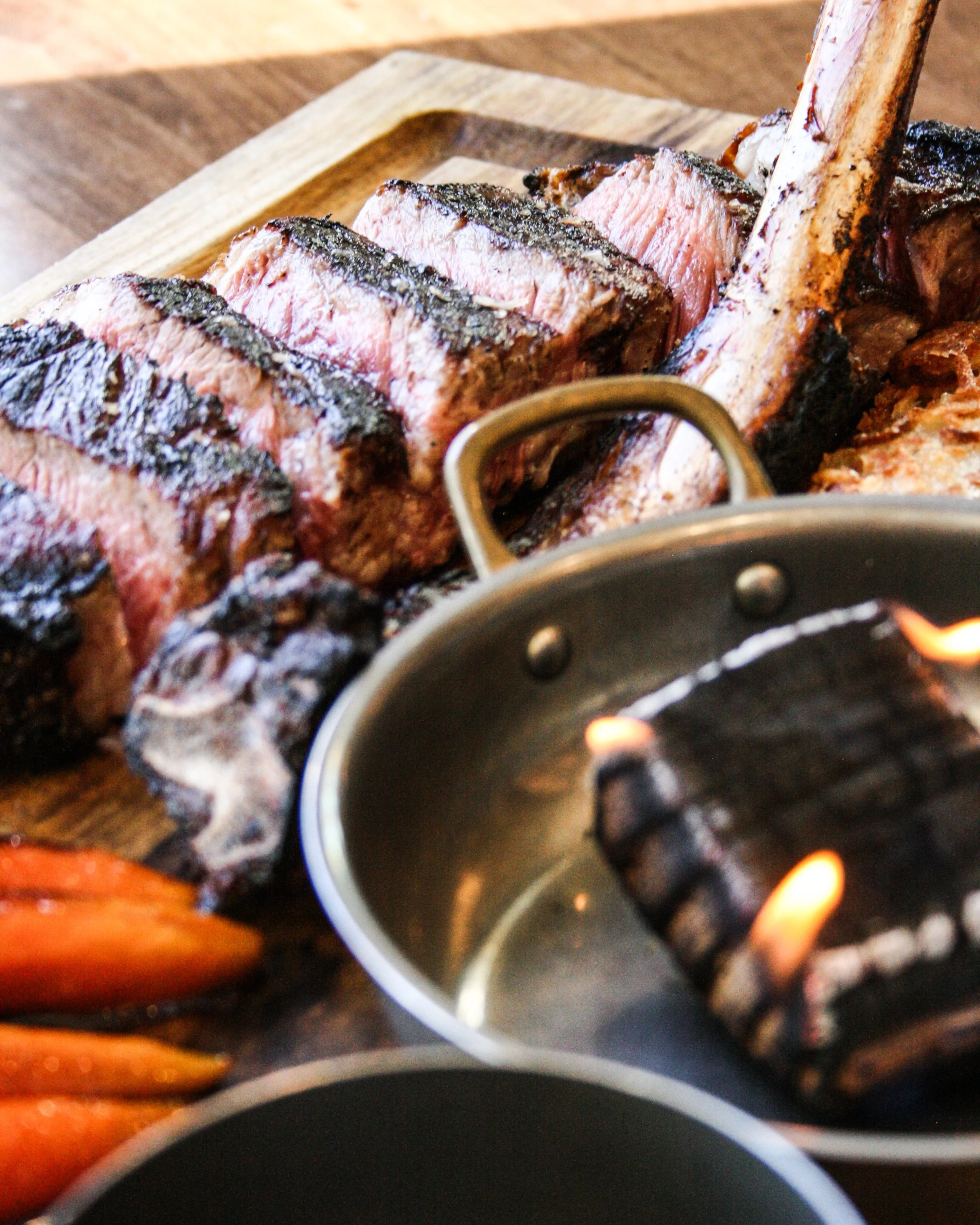 Raw steak on a wooden cutting board, with a small pan of flame and a piece of lit wood nearby.