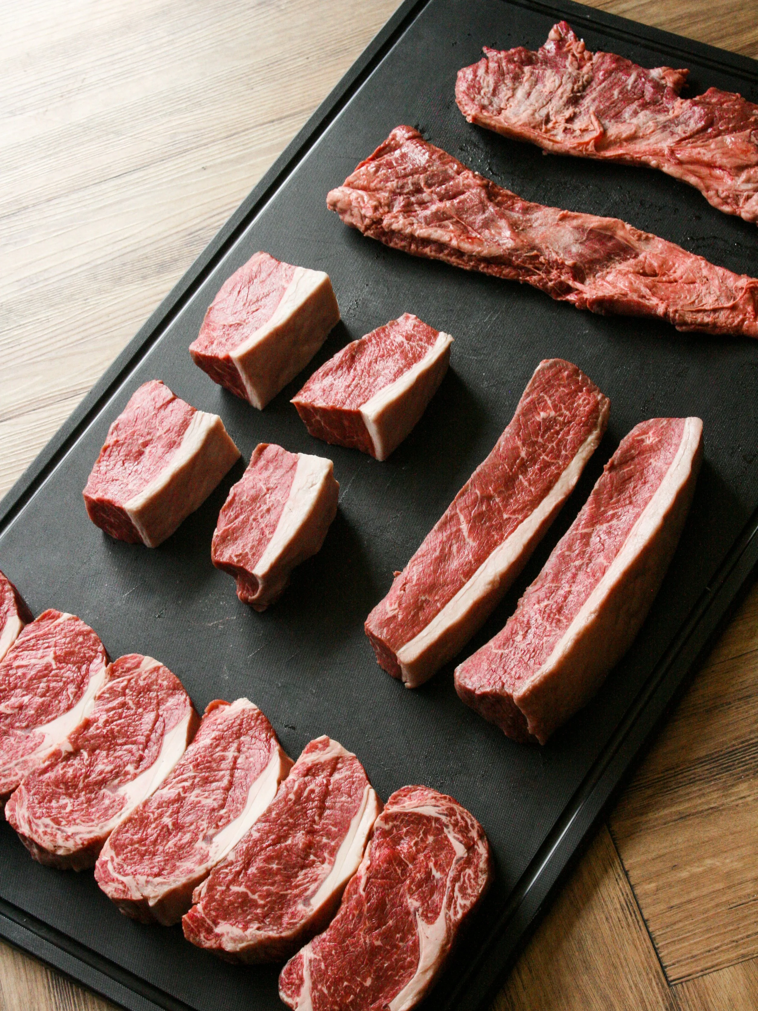 Assorted cuts of raw beef on a black cutting board, including steaks and strips, on a wooden surface.