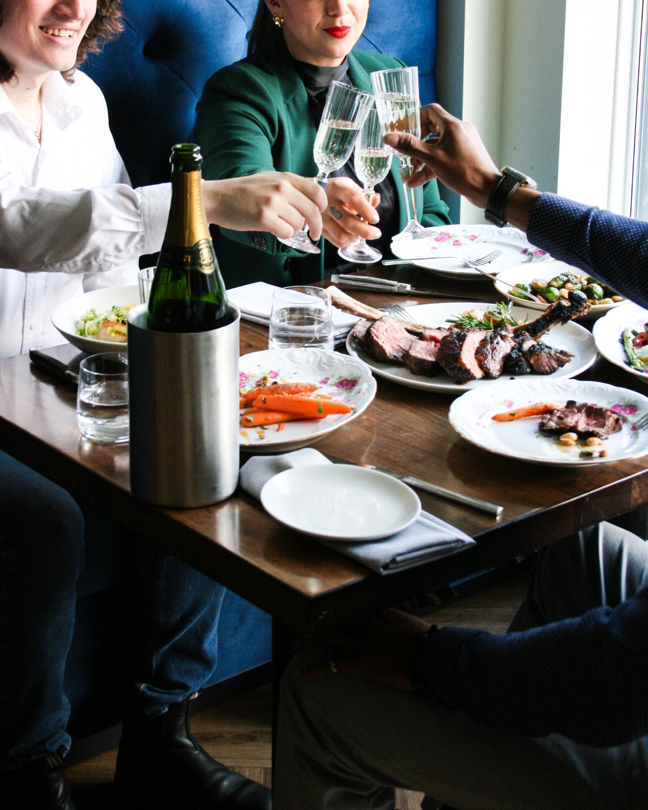 People gathered around a dining table sharing a meal, raising glasses of champagne for a toast, with various plates of food including steak and vegetables on the table.