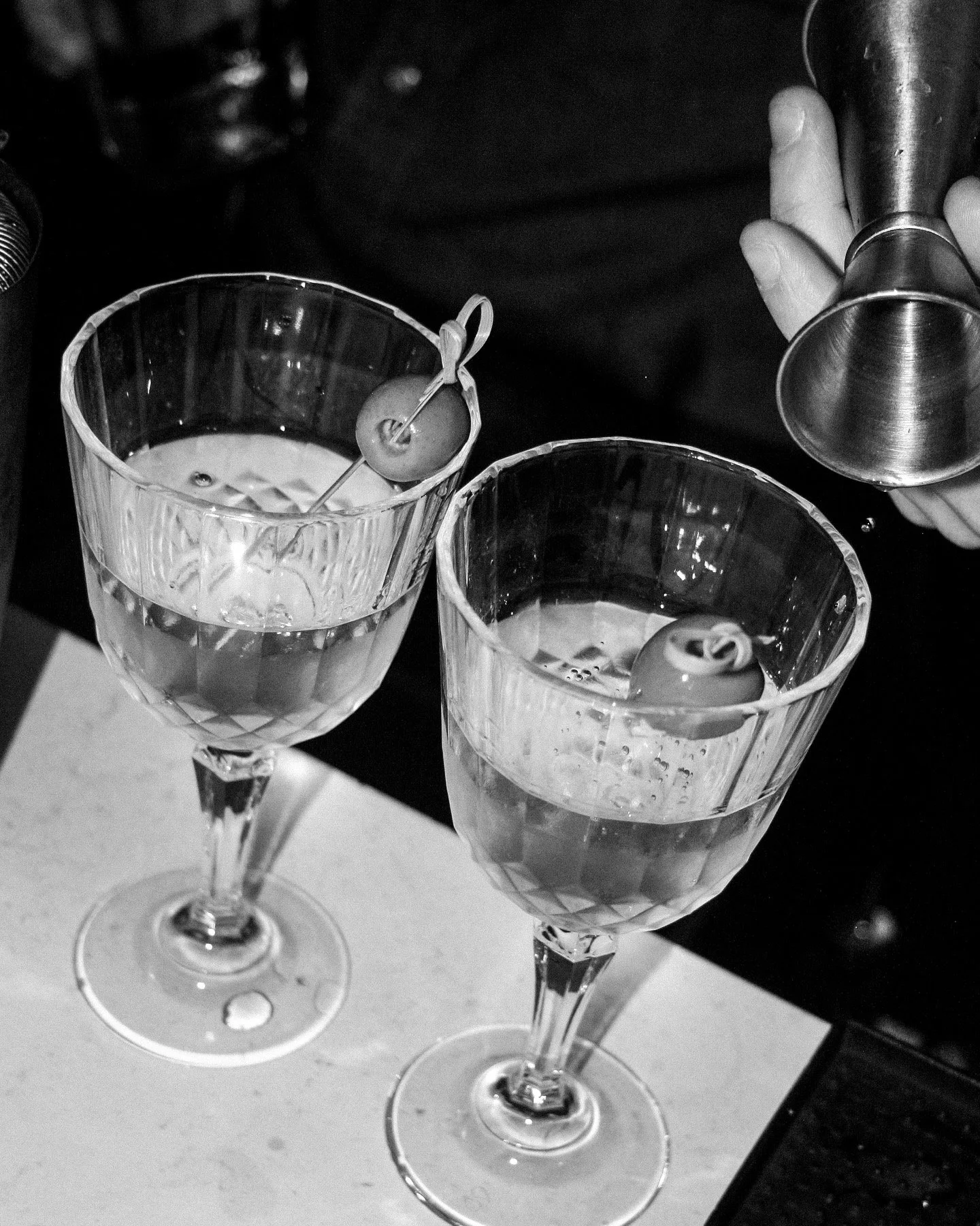 Two crystal glasses filled with a clear liquid, garnished with cherries, and an alcoholic drink being poured into one of the glasses.