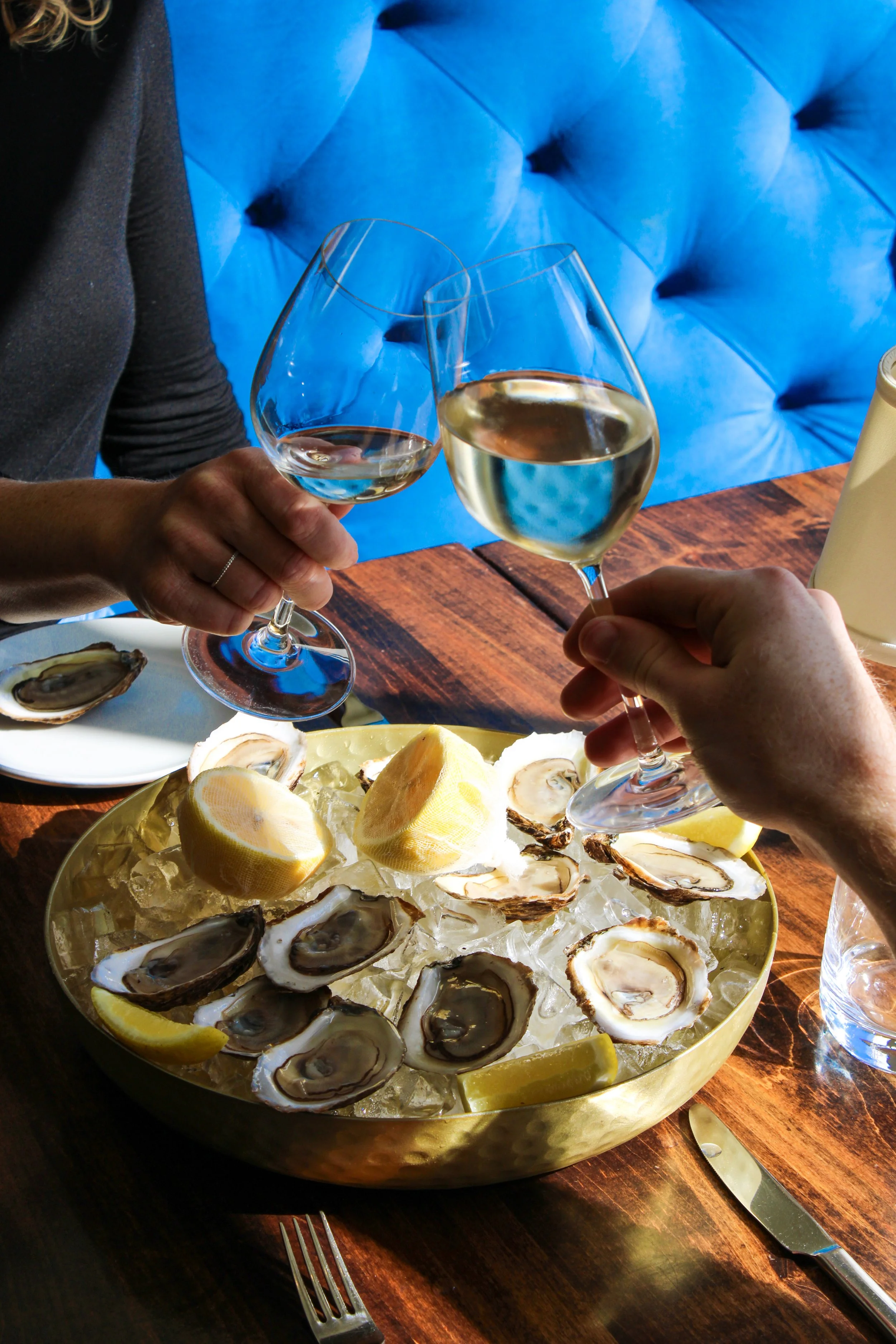 Two people clinking glasses of white wine over a bowl of oysters on a bed of ice with lemon wedges.