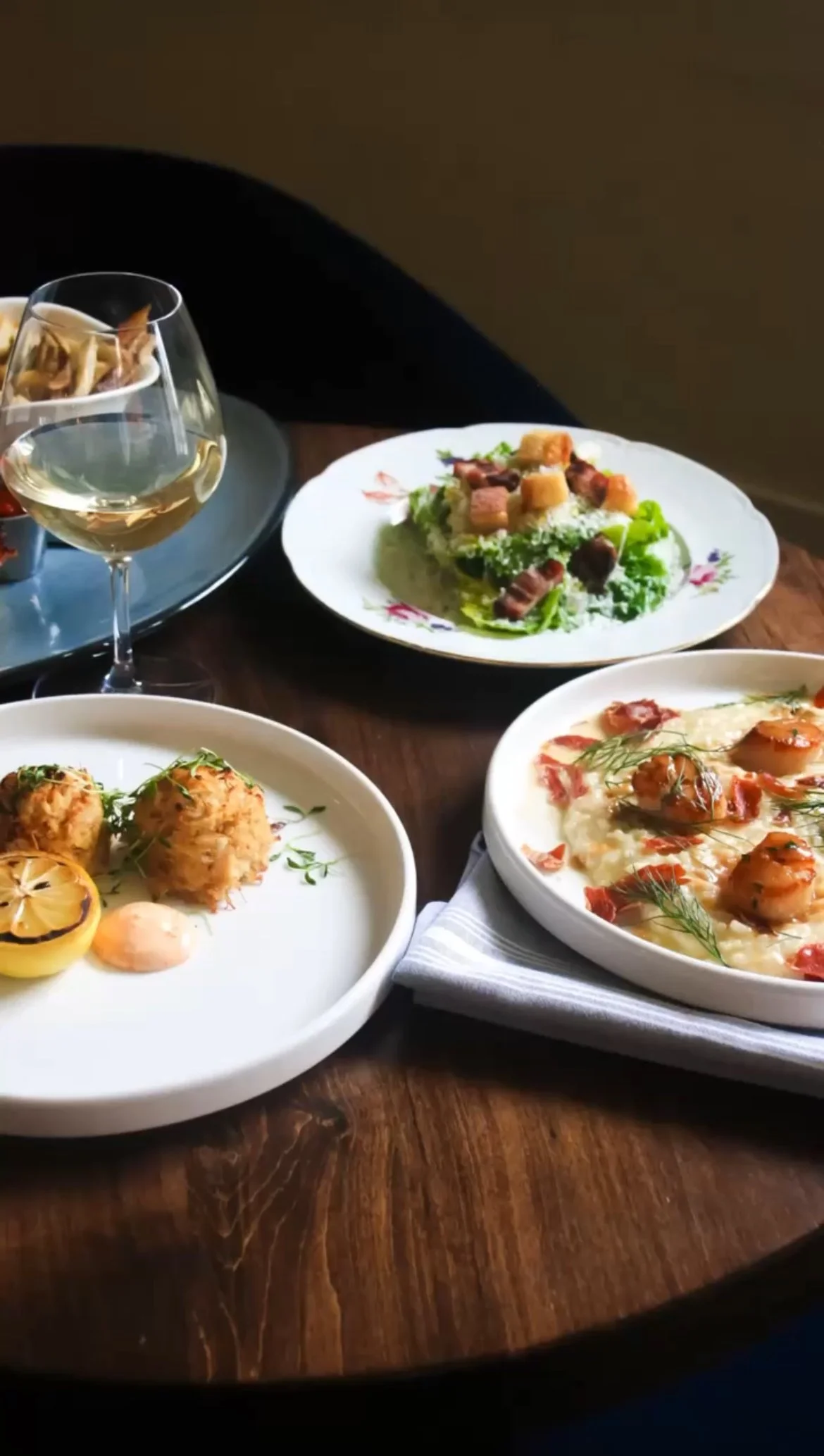 A table with plates of gourmet food including crab cakes, scallops, risotto, a classic Caesar salad, in moody restaurant lighting.