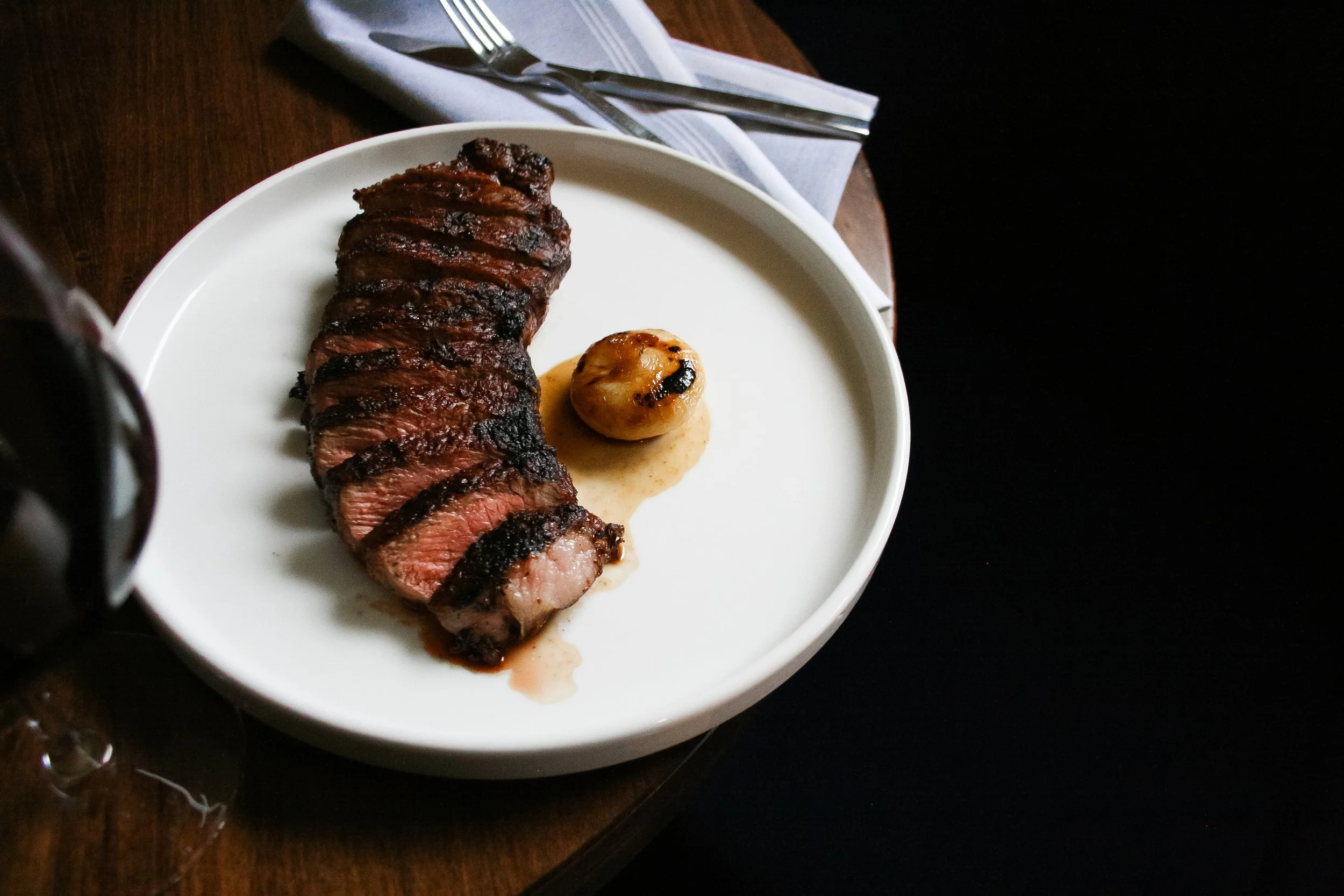 A medium-rare steak sliced and grilled with grill marks, served on a white plate with a roasted garlic clove and jus, on a dark wooden table with utensils on a napkin.