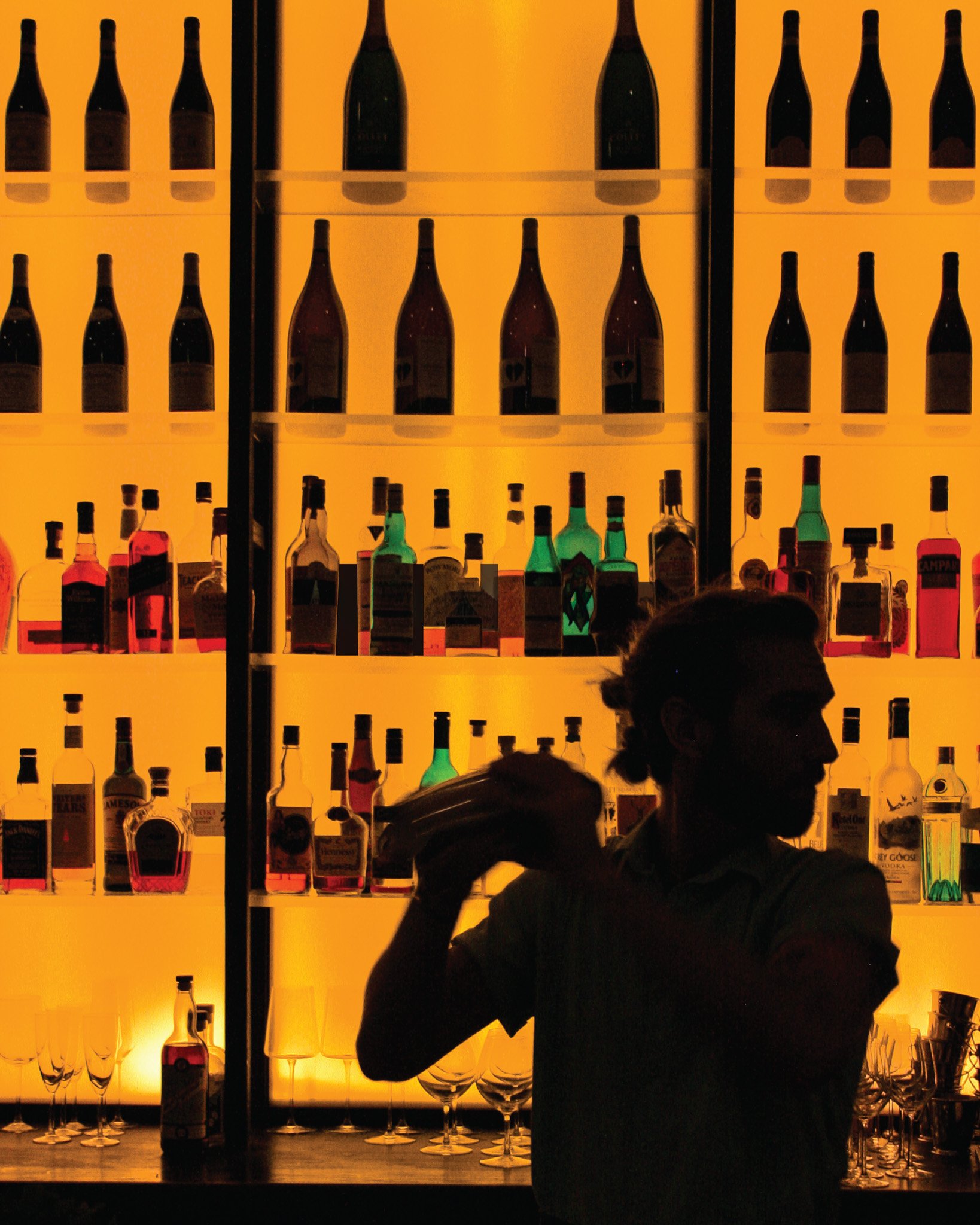 Silhouette of a bartender holding a cocktail shaker in front of a brightly lit bar with shelves of bottles