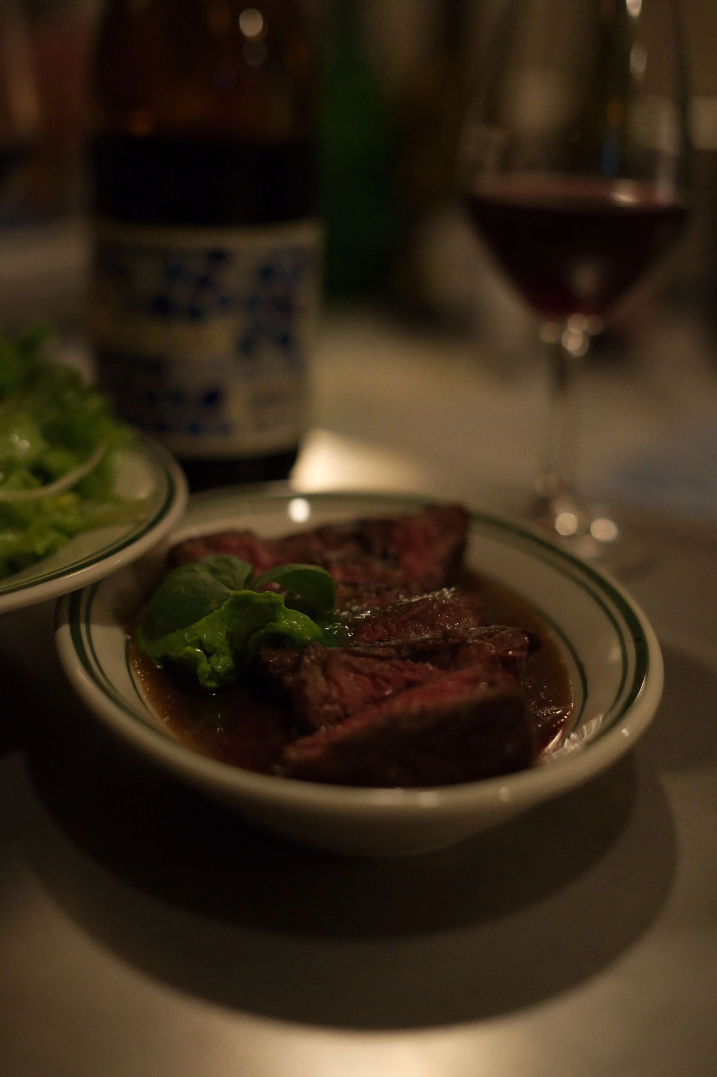A bowl of cooked steak with green garnish, a glass of red wine, and a container of water on a dimly lit table