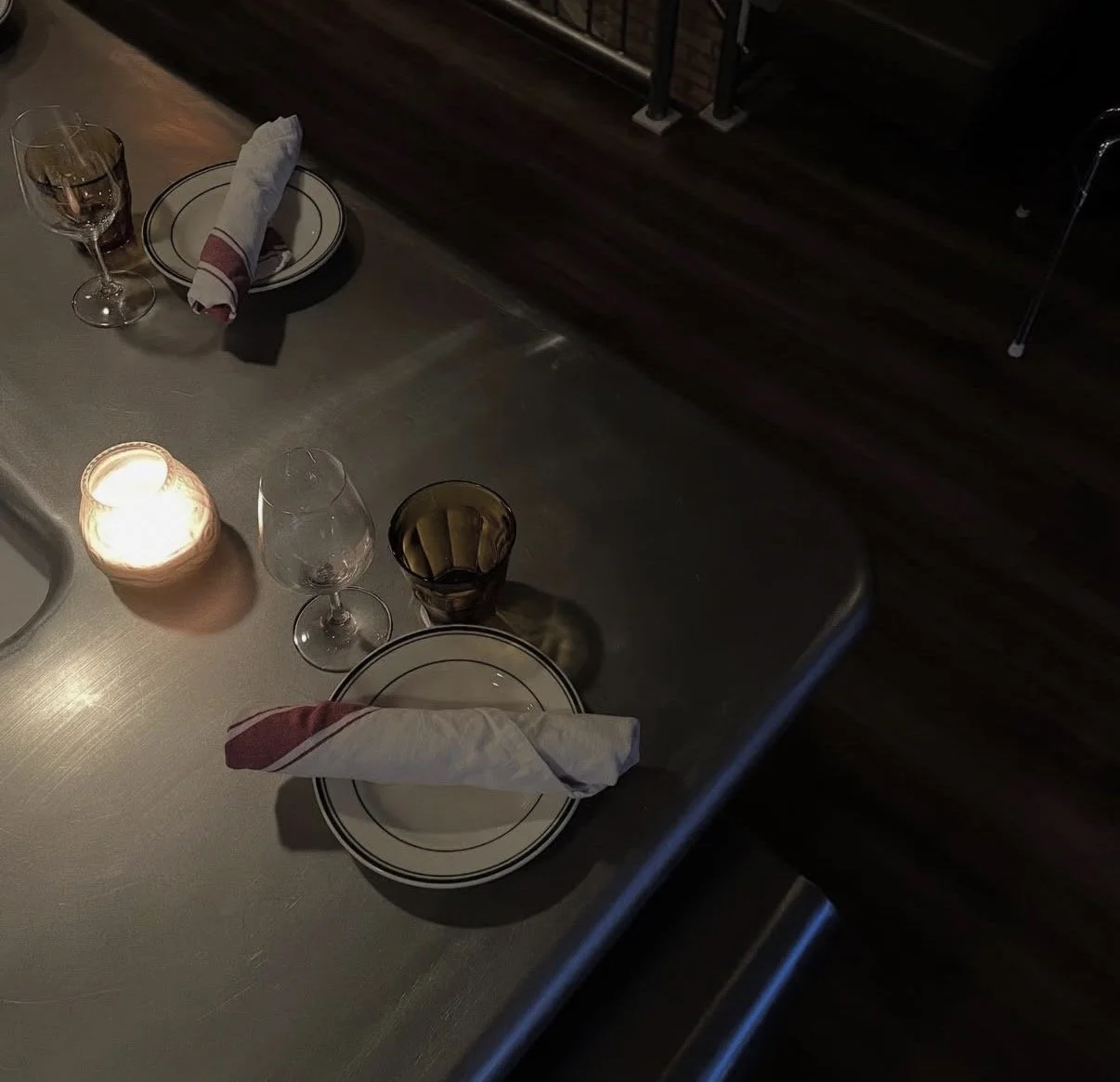 A dimly lit dining table set for two with two plates, each with a folded cloth napkin, a wine glass, a water glass, and a candle in a jar providing warm light.