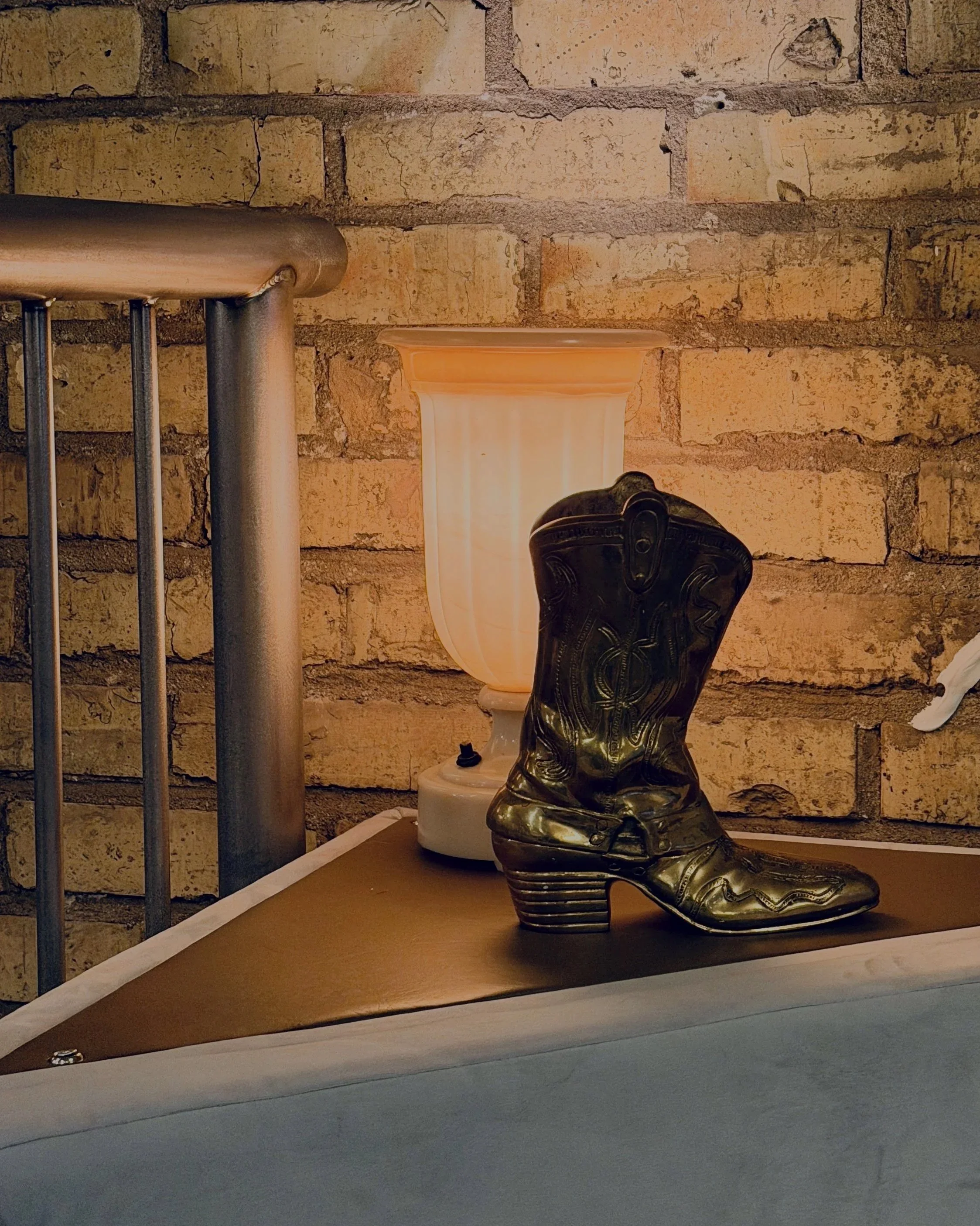 A decorative gold cowboy boot sculpture on a table, with a beige lamp and a brick wall in the background.