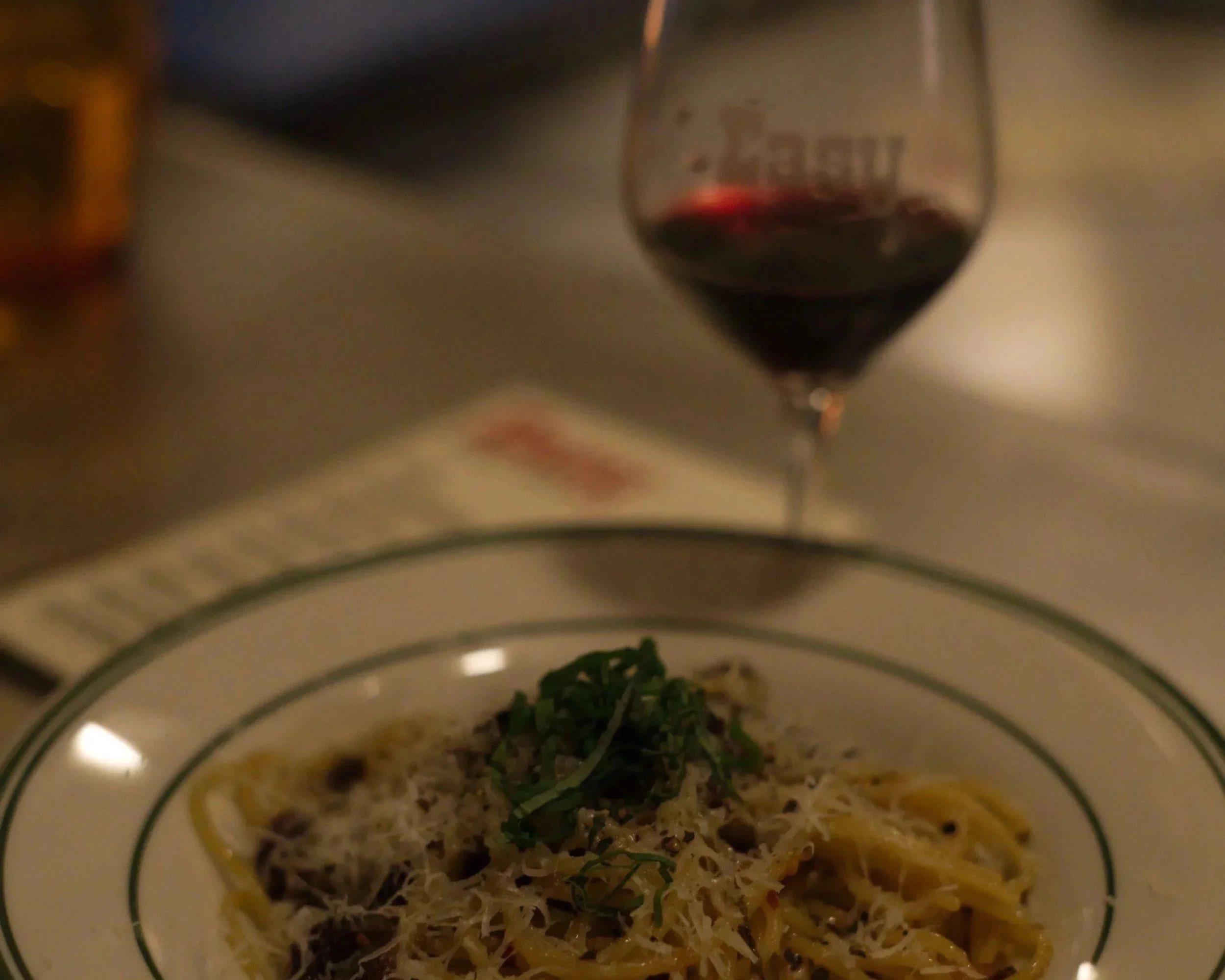 A plate of pasta topped with grated cheese and herbs, with a glass of red wine in the background.