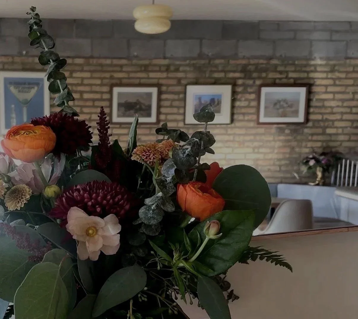 Close-up of a colorful flower bouquet with orange, burgundy, pink, and cream flowers and green foliage, inside a room with brick walls, framed pictures, and dining tables in the background.