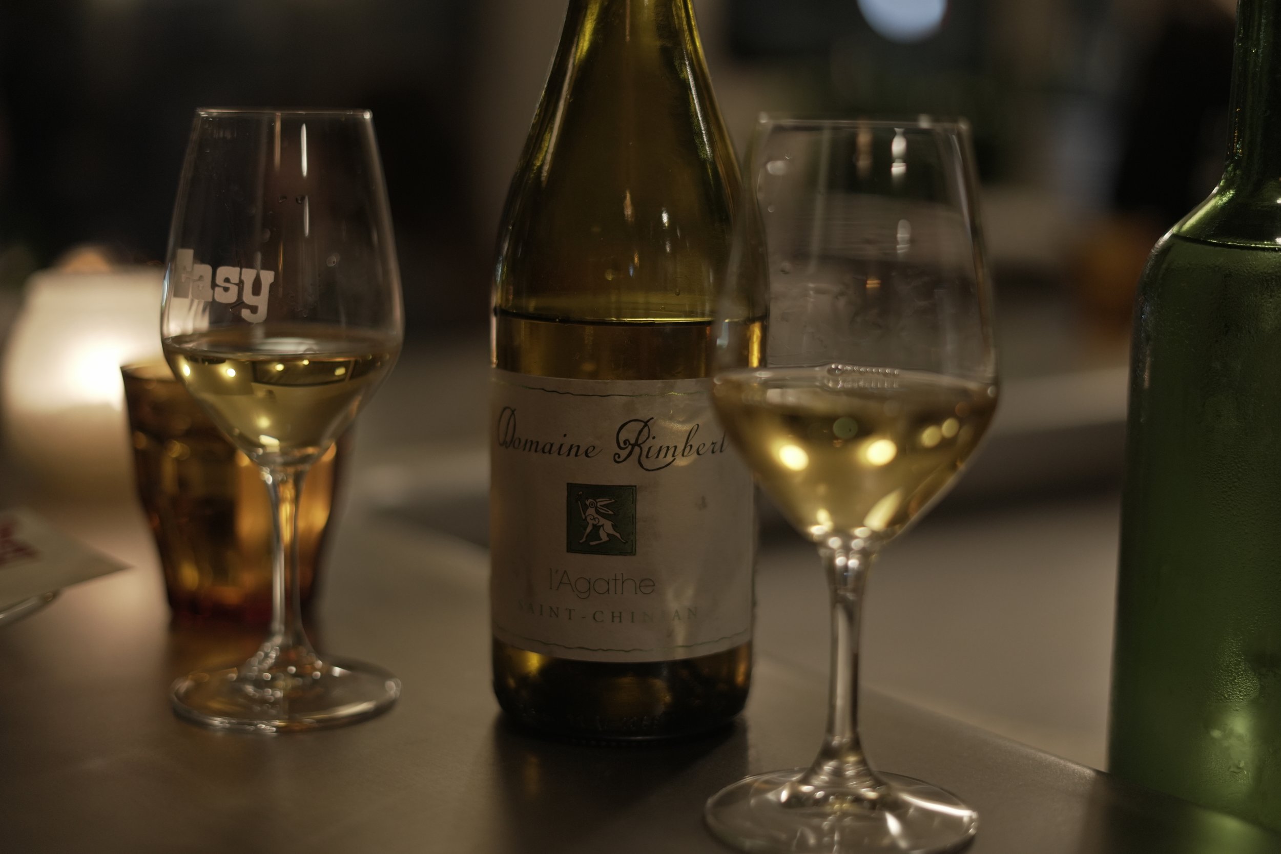 Two glasses of white wine, a bottle of Domaine Kimberl wine, and a green bottle on a table in a dimly lit setting, possibly at a dinner or gathering.