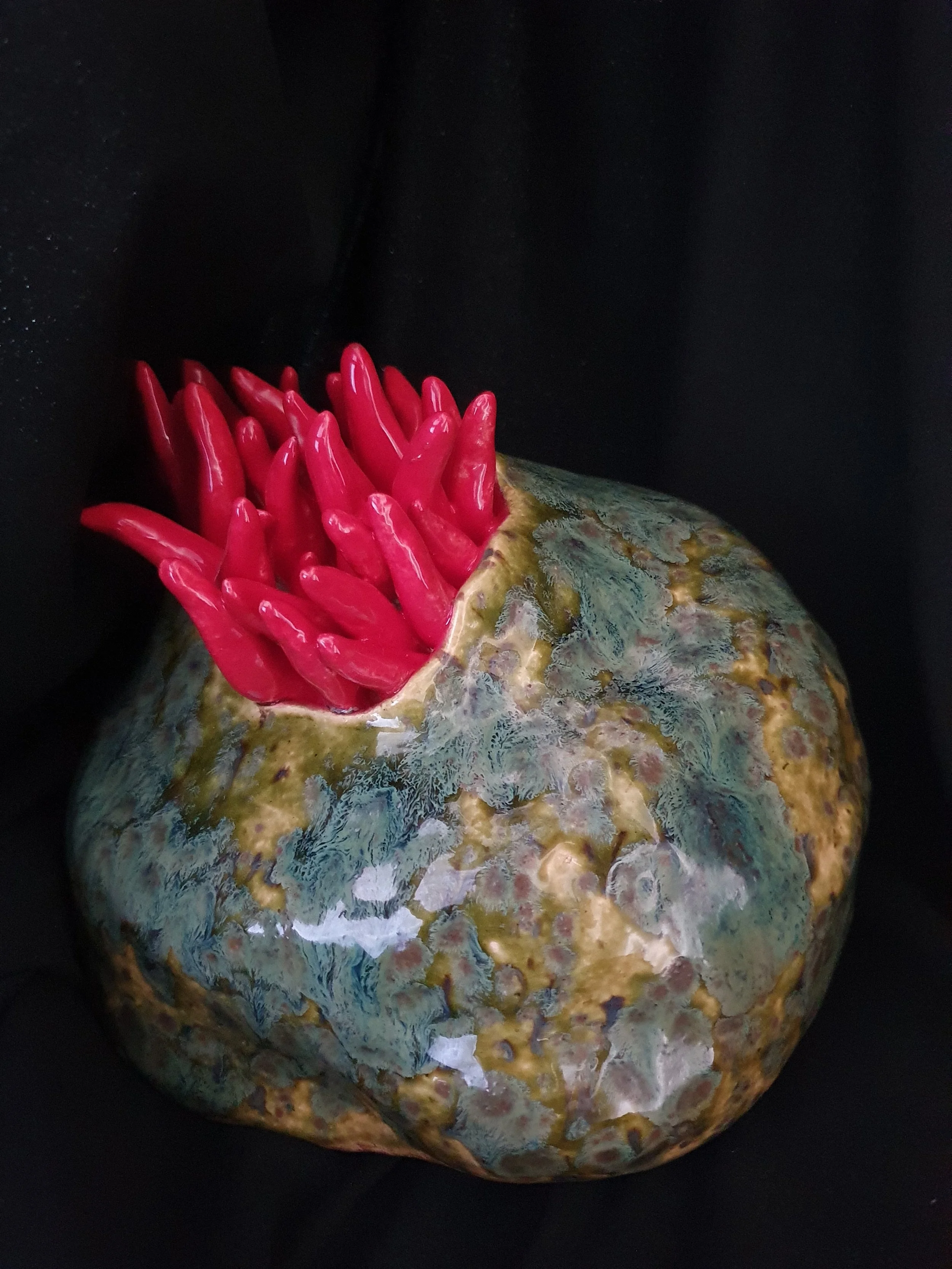 An unusual sculpture of a pomegranate with a glossy, red interior resembling chili peppers, set against a black background.
