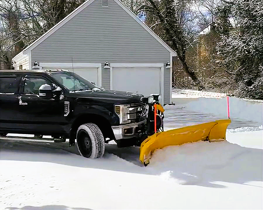 Black pickup truck with a yellow snow plow attached to its front, parked on snow-covered ground in front of a gray garage with white doors, in a snowy residential area with trees in the background.