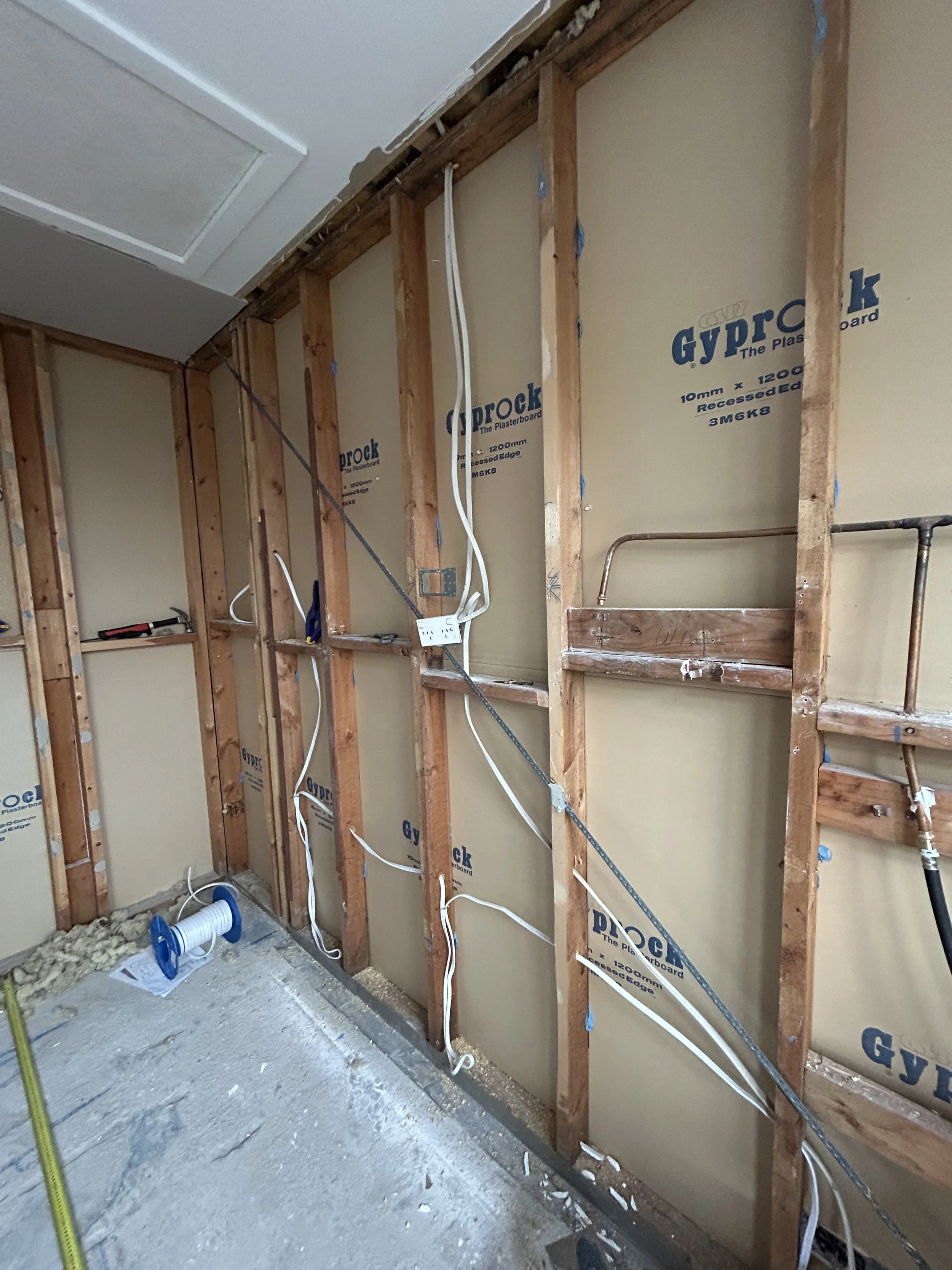 Interior view of a wall under construction with exposed wooden wall studs, electrical wiring, and ceramic pipe. Insulation and drywall sheets are partially installed.