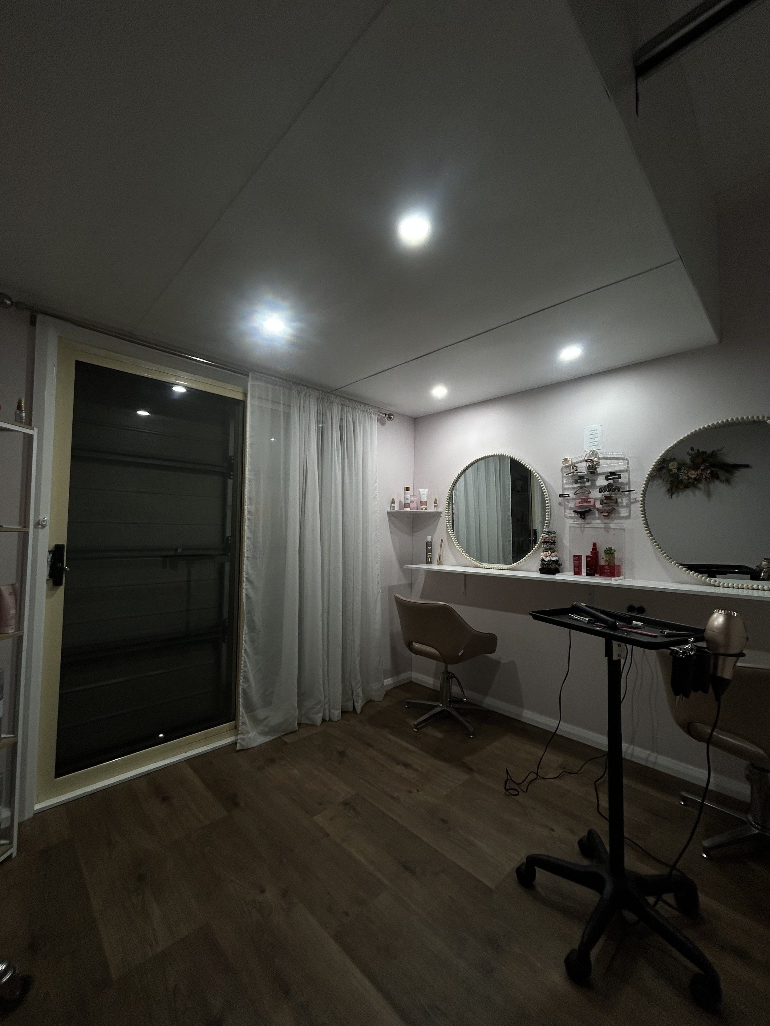 A hair salon with two mirrors, two chairs, and a styling station. The room features hardwood flooring, white walls, and ceiling lights. There's a window with white curtains and various hair products and tools on the shelves.