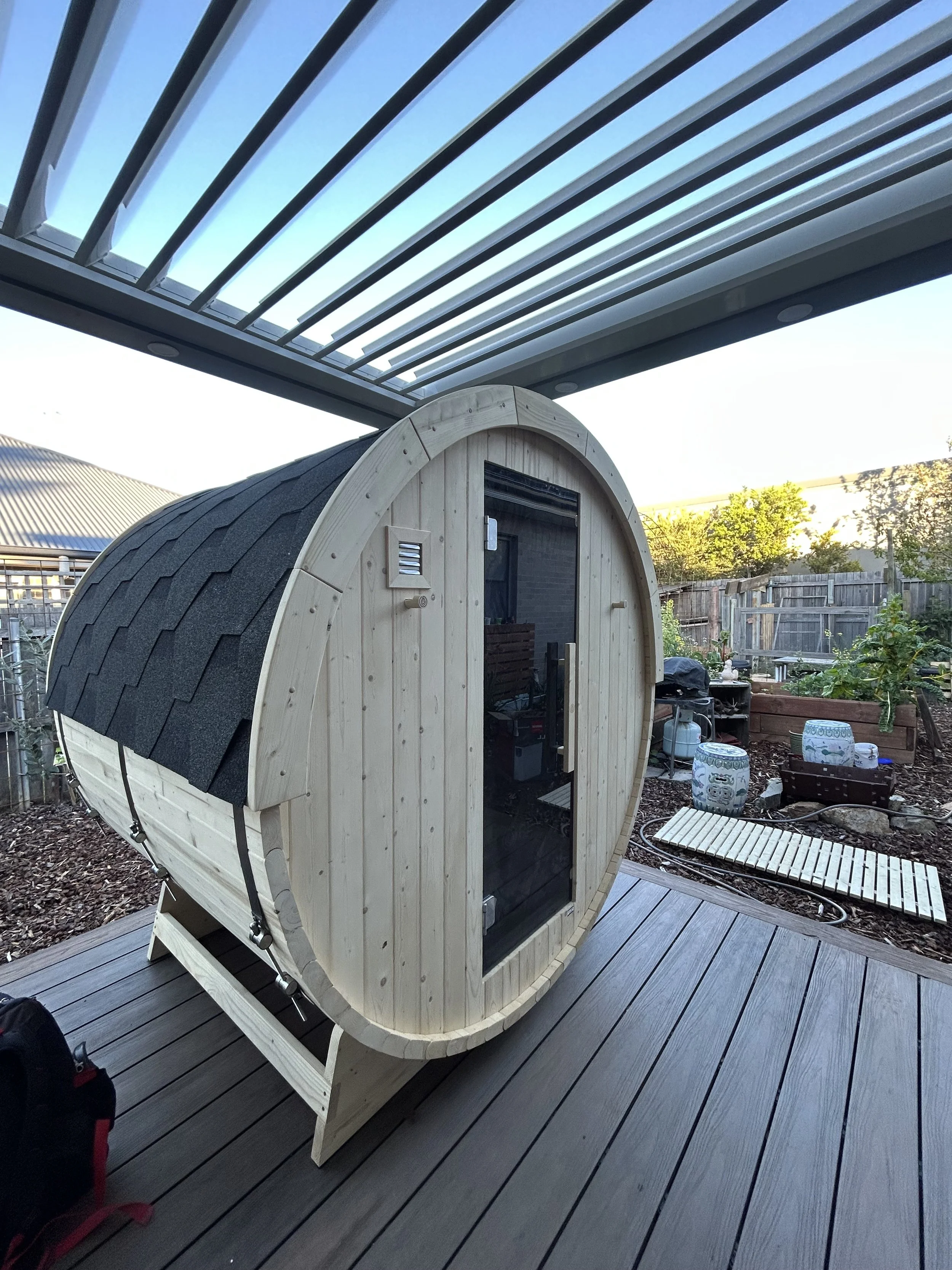 A wooden barrel-shaped outdoor sauna with a glass door, situated on a wooden deck in a backyard garden.