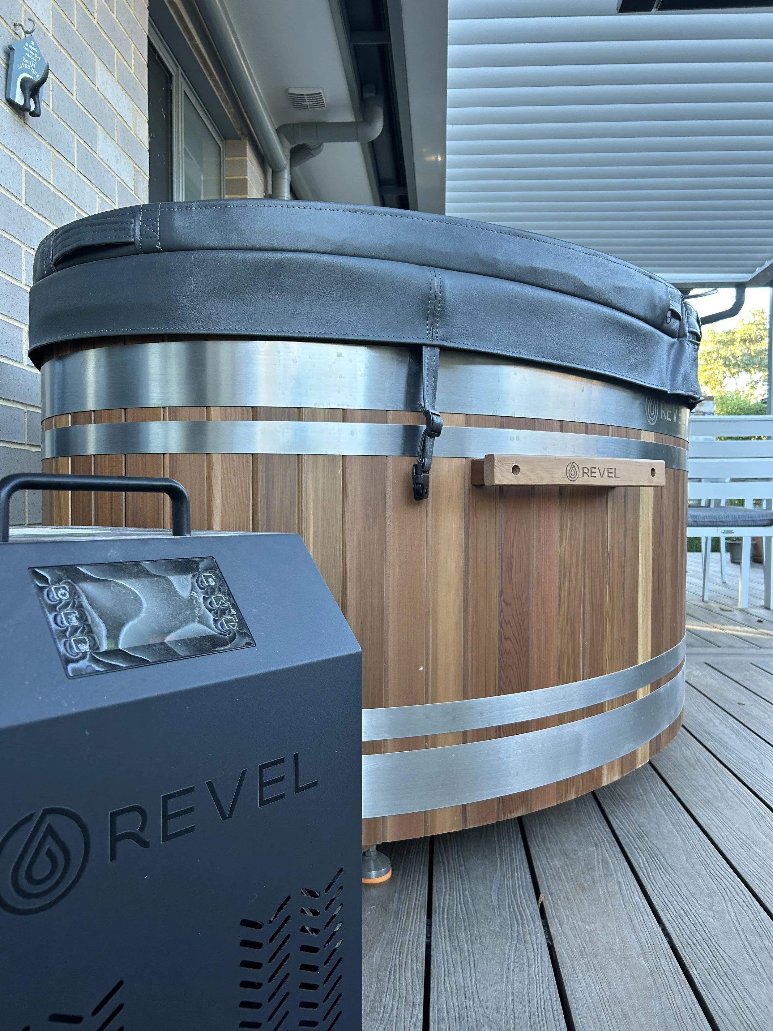A wooden hot tub with a padded cover on the top, situated on a wooden deck outdoor area. A black REVEL lounge chair or equipment is partially visible in the foreground.