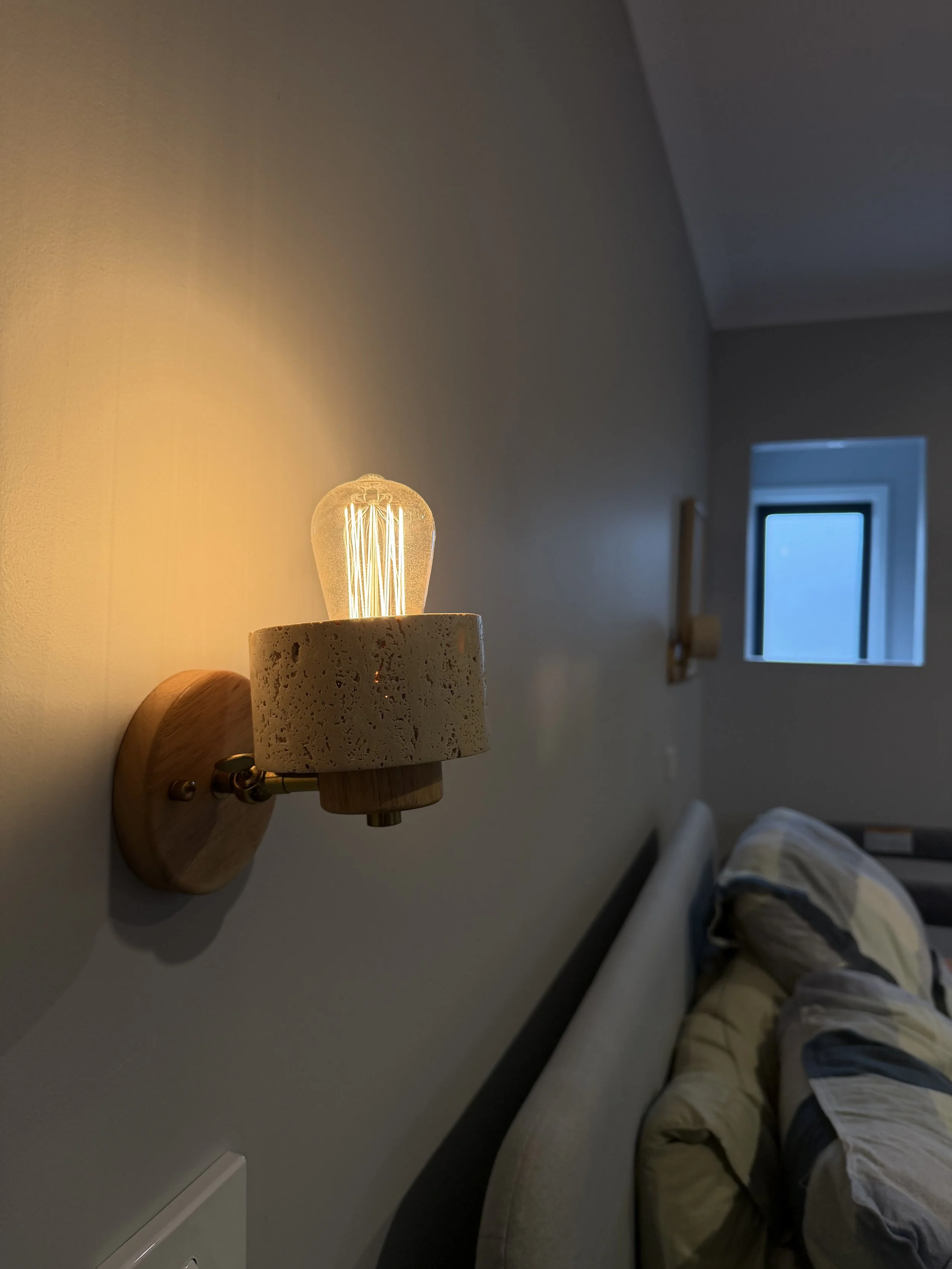 Wall-mounted bedside lamp with a warm glowing filament bulb, above a bed with a patterned comforter, in a bedroom near a window.
