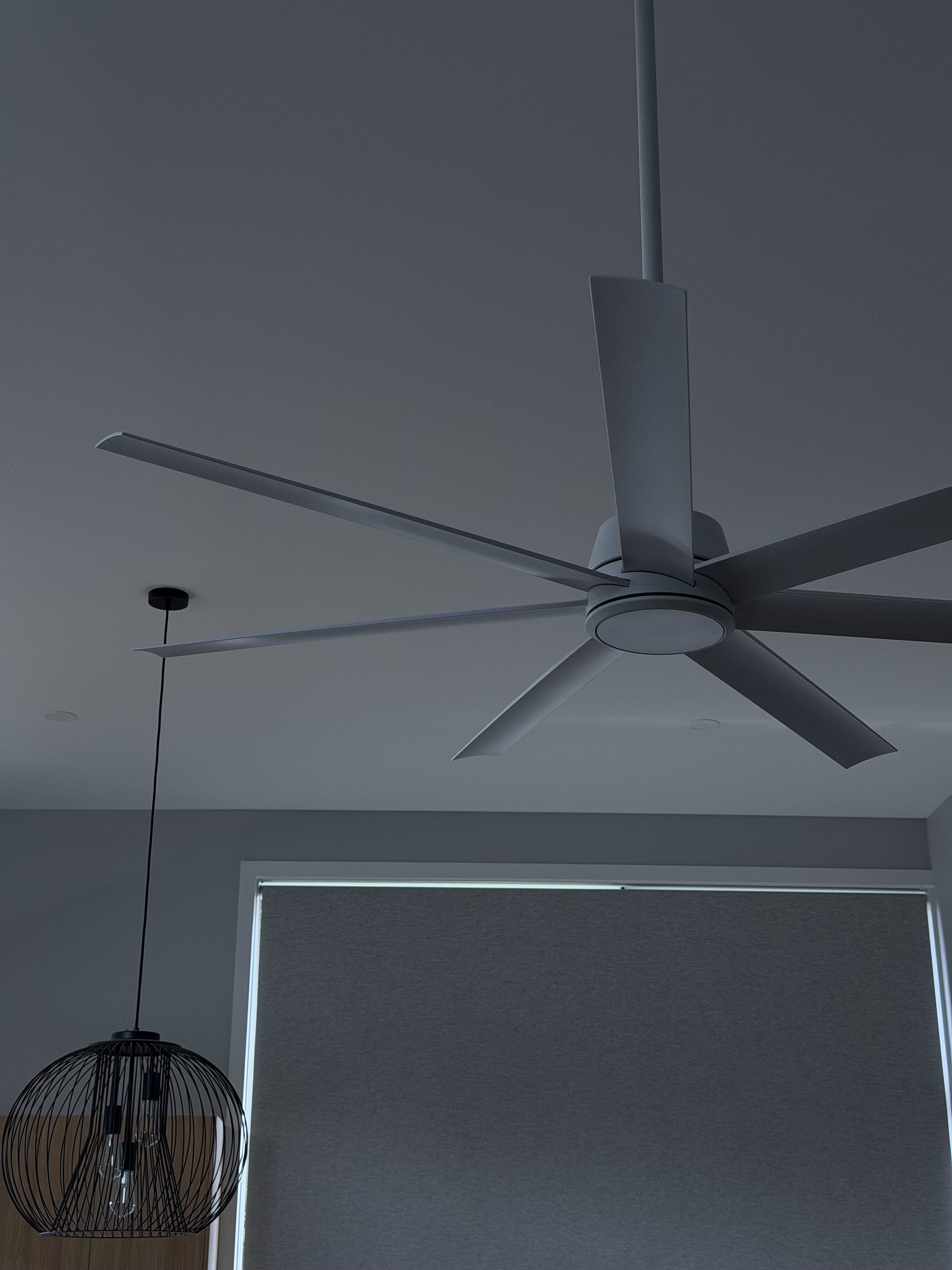 A modern ceiling fan with five blades and a ceiling lamp with a black wire cage design in a room with gray walls and a window covered by a gray roller shade.