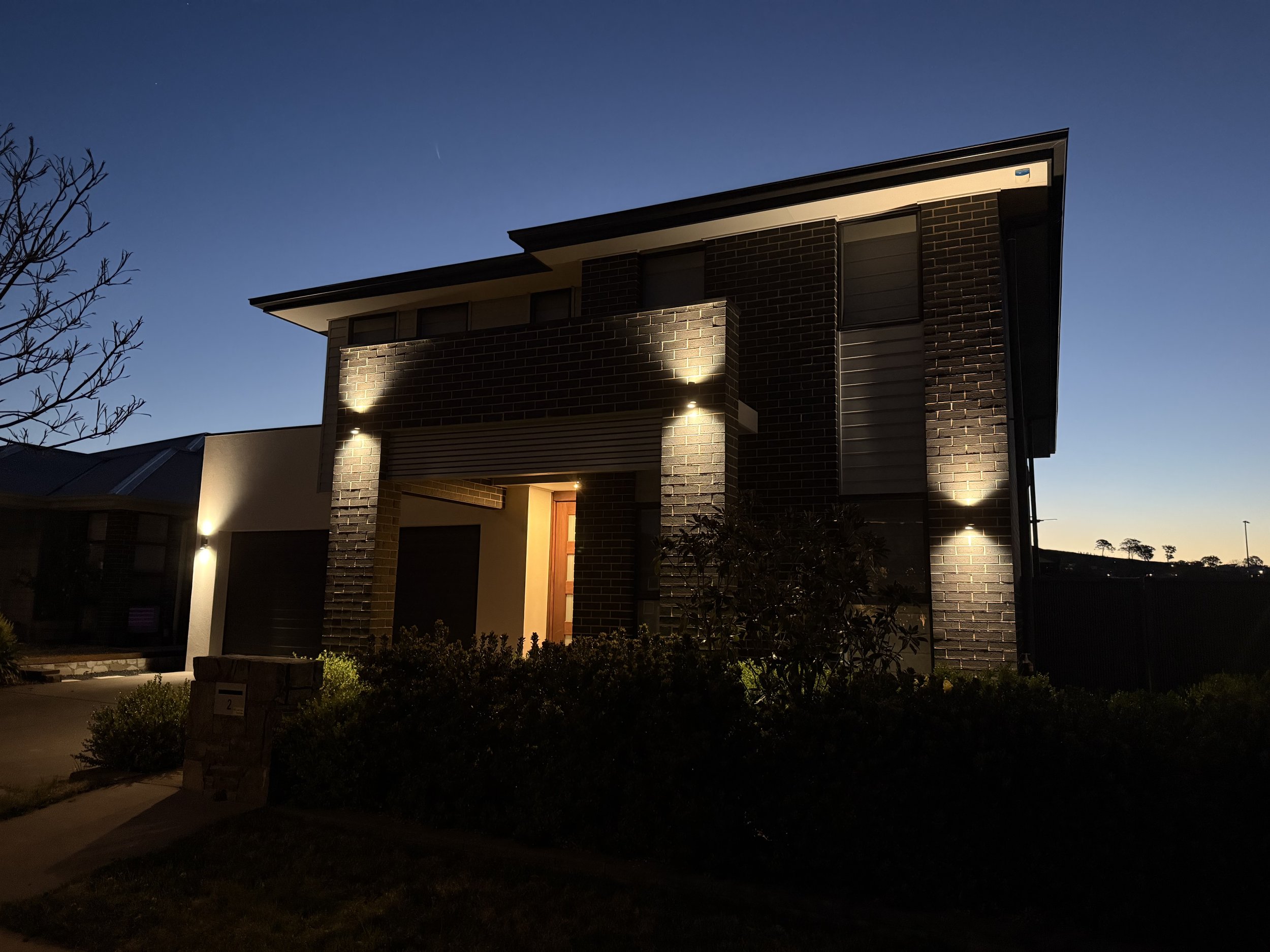 A modern two-story house at dusk, illuminated by exterior lights, with a brick facade, large windows, and surrounding shrubs.