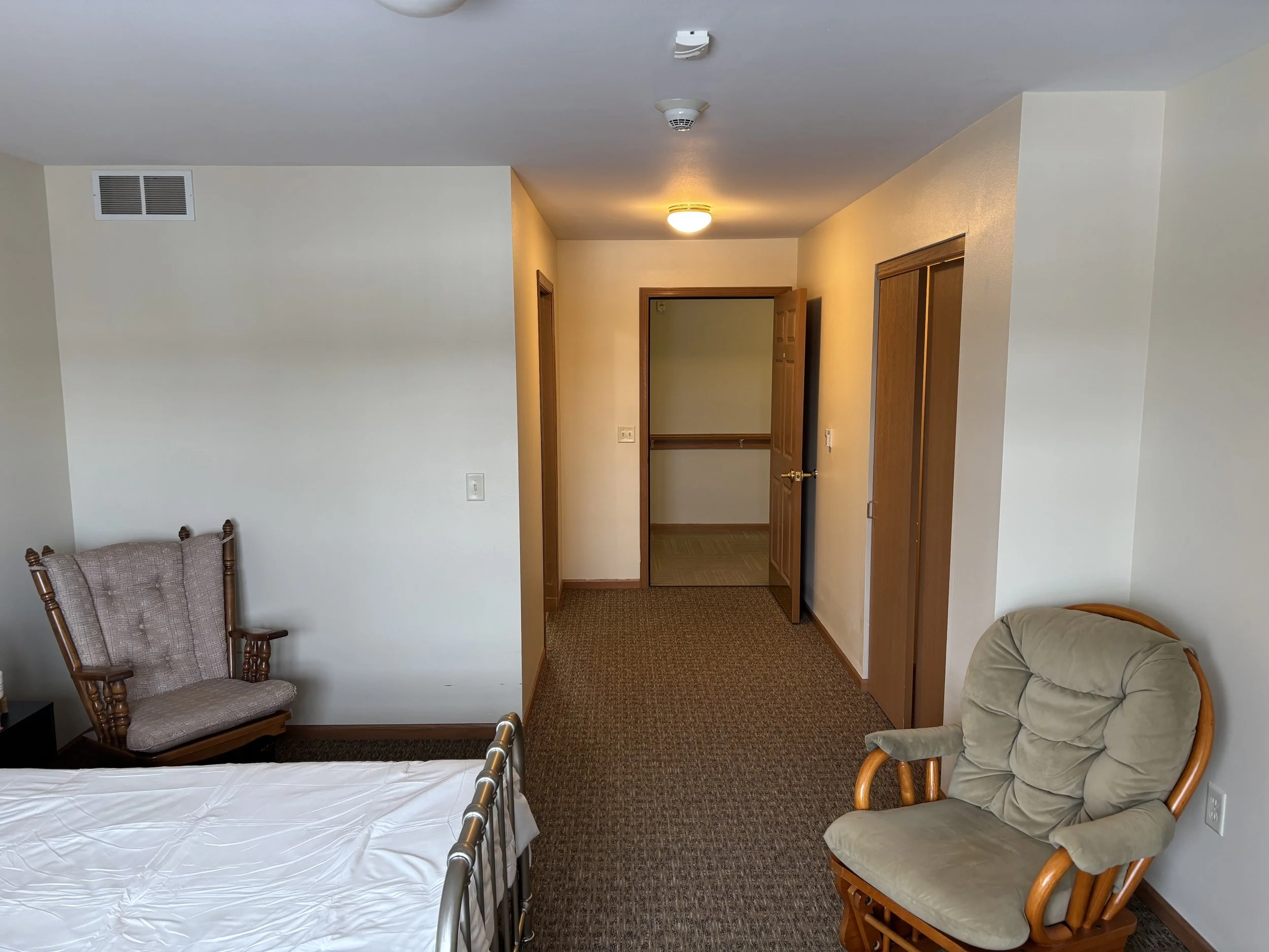 A bedroom with a bed, two chairs, and a hallway with wooden doors.