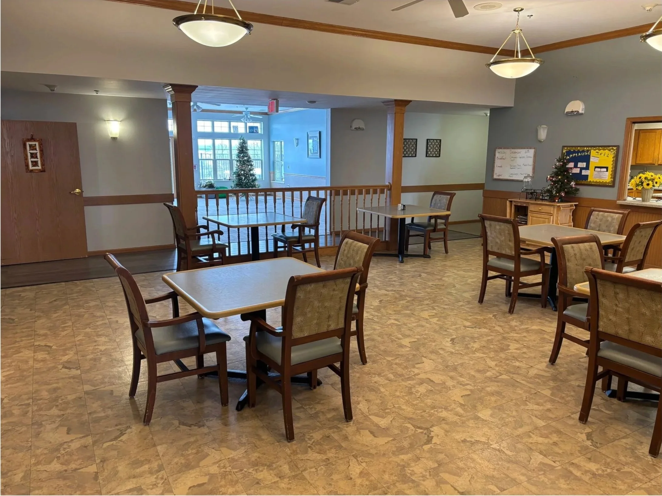Indoor dining area with several tables and chairs, decorated for Christmas with a small Christmas tree and a sunflower arrangement, and featuring a large window with a Christmas tree outside.