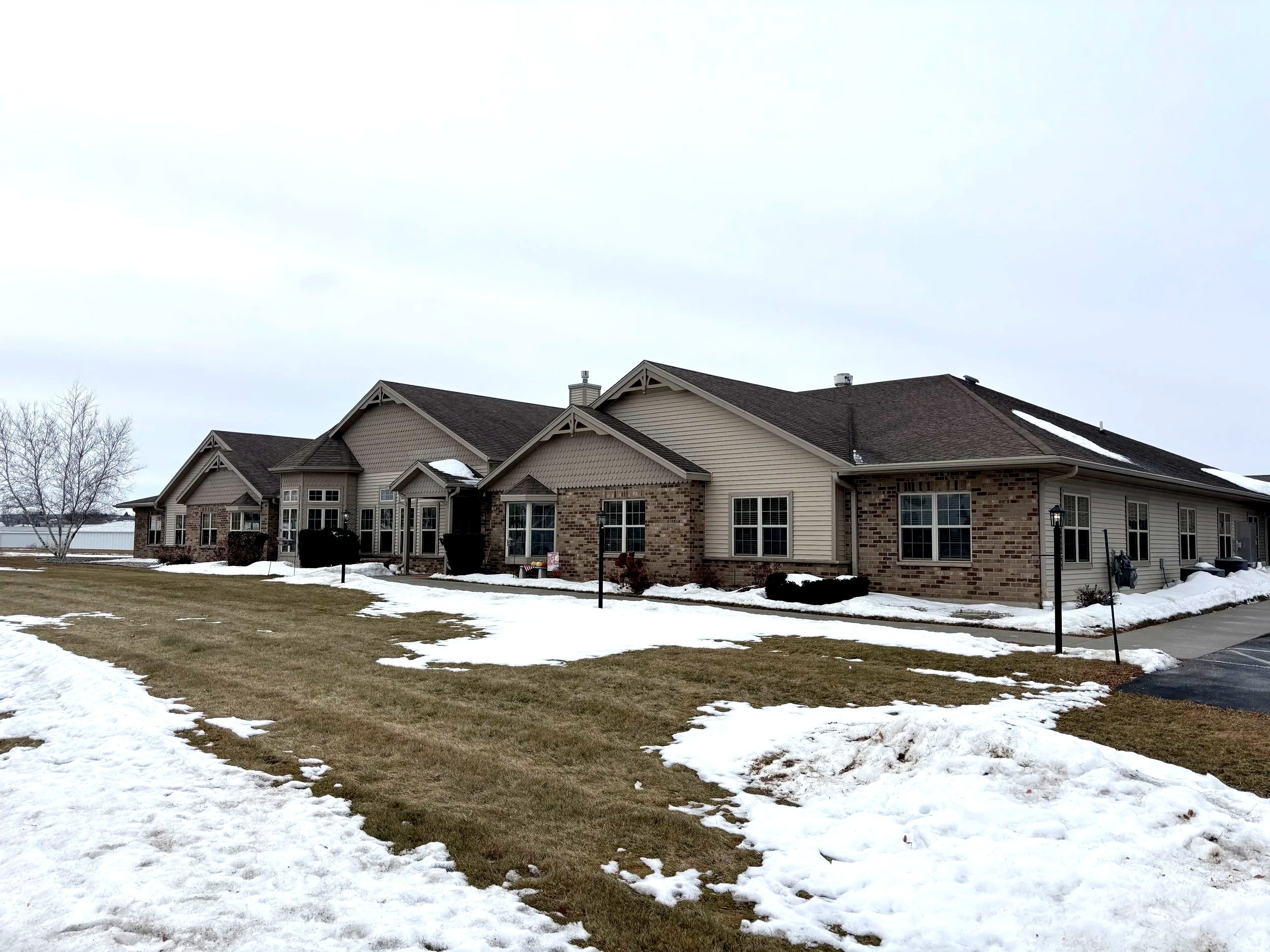 Large house with brick and siding exterior, snow on the ground, some patches of grass visible, overcast sky.