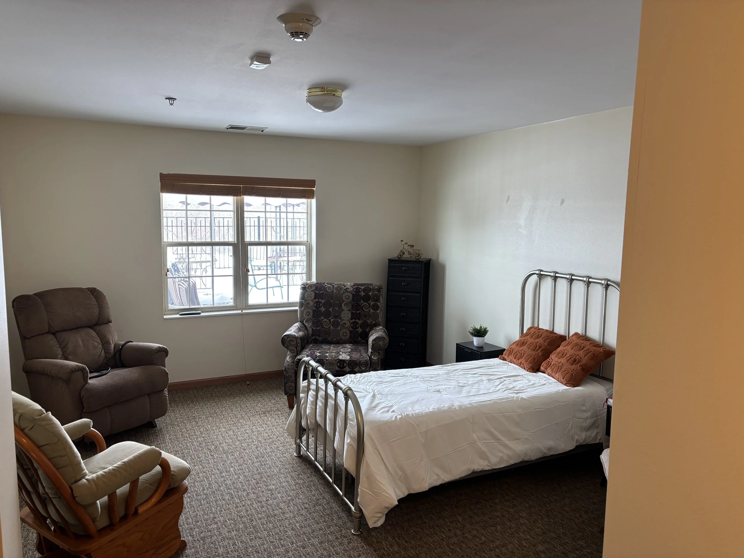 A bedroom with a bed, two orange pillows, a small black side table with a plant, a black dresser with a small decorative bicycle, a patterned armchair, a recliner, a wooden rocking chair with white cushions, and a window with blinds