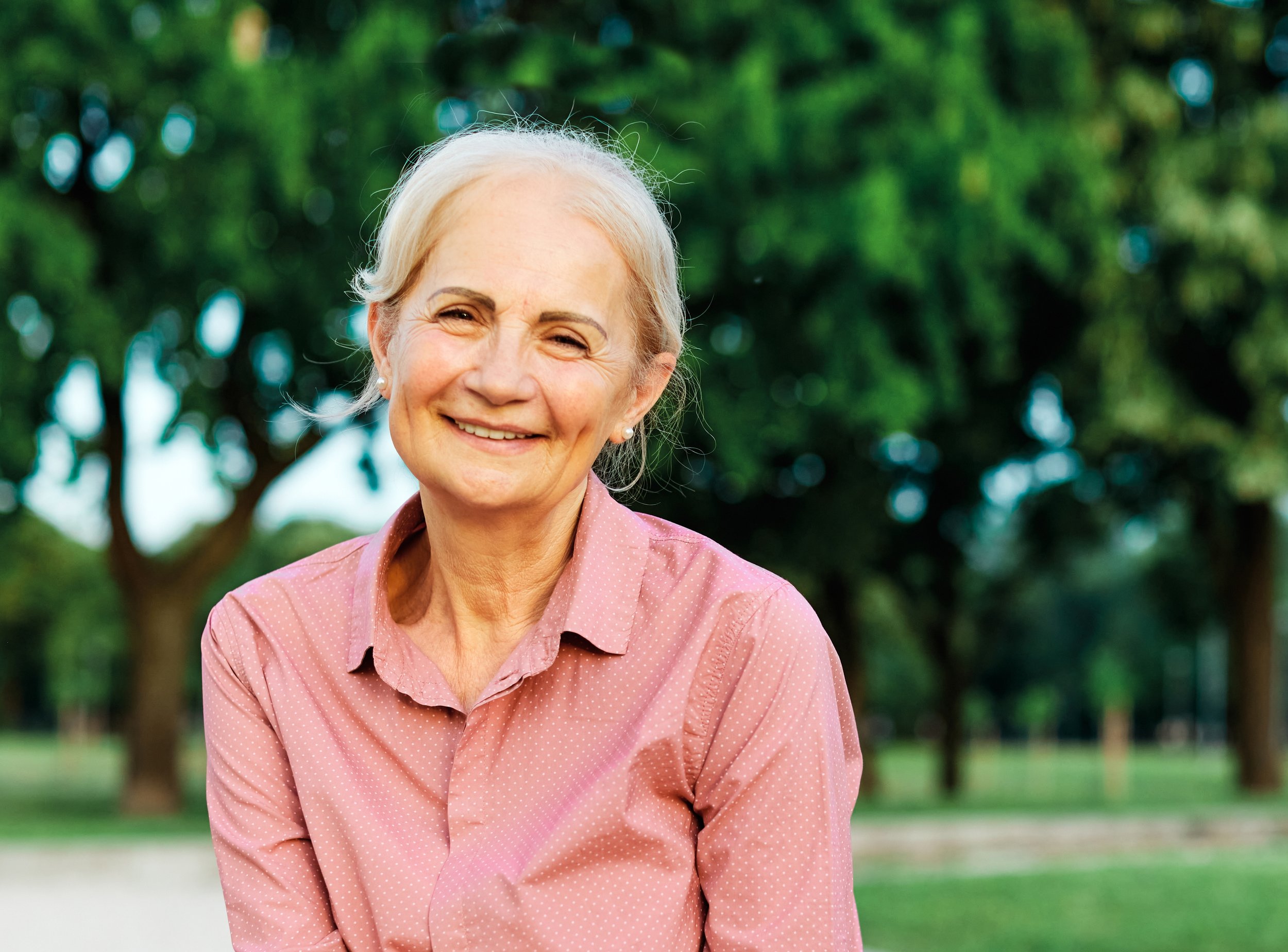 An elderly woman with gray hair smiling outdoors, wearing a pink polka-dotted shirt, with green trees in the background.