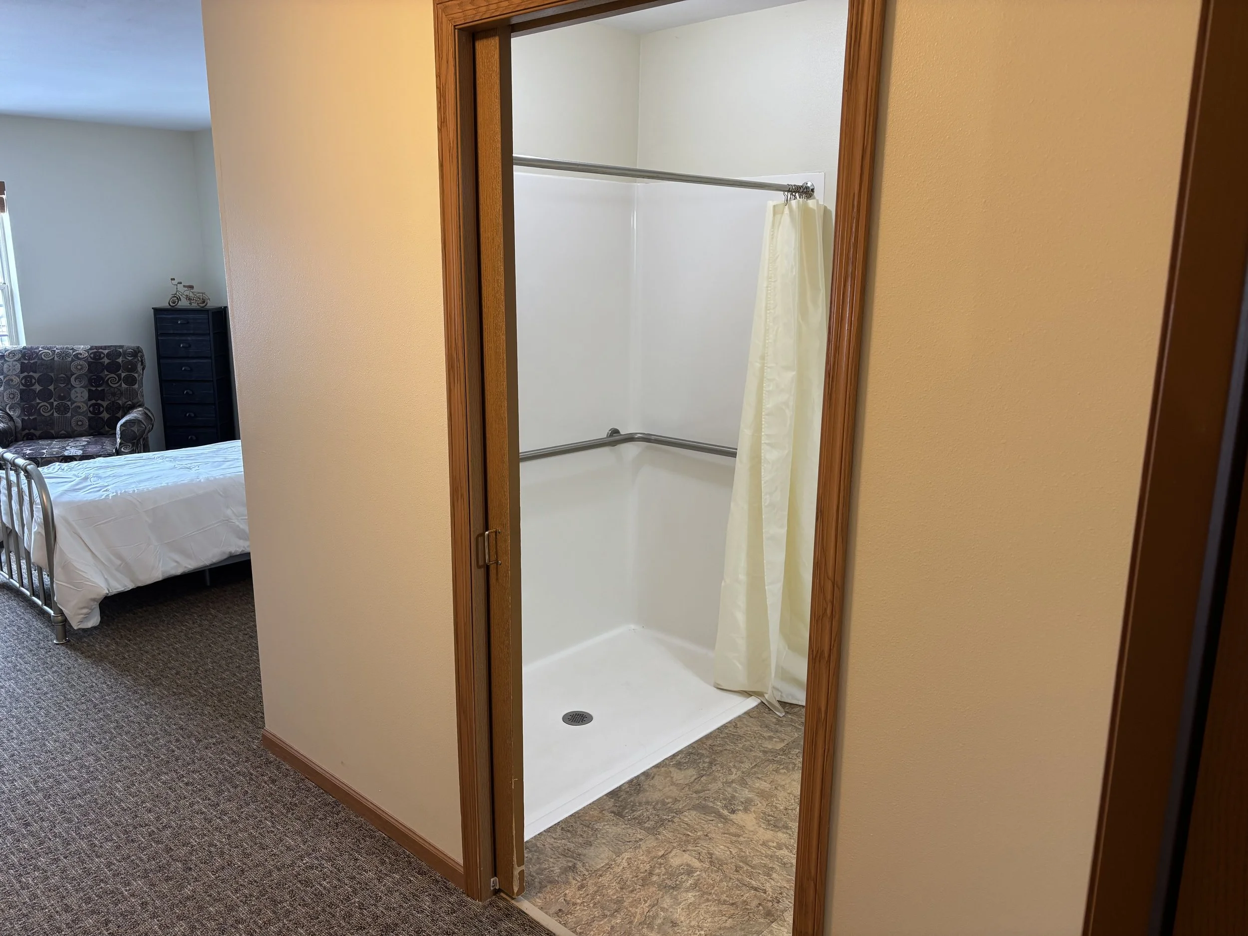 Inside a room showing a bathroom with a walk-in shower with a cream-colored shower curtain, and a bedroom with a bed, armchair, and dresser in the background.